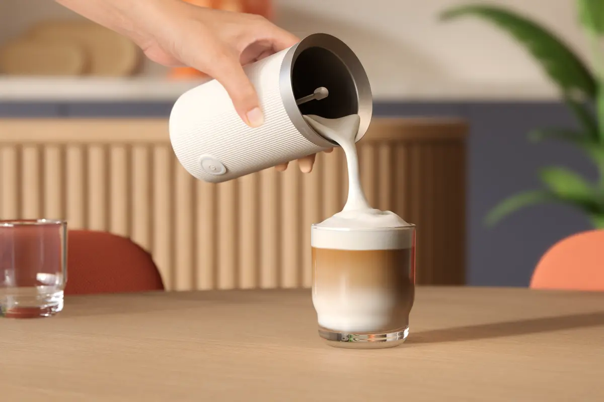 Una mano versa latte schiumoso in una bevanda al caffè a strati. Un montalatte bianco è tenuto sopra un bicchiere su un tavolo di legno.