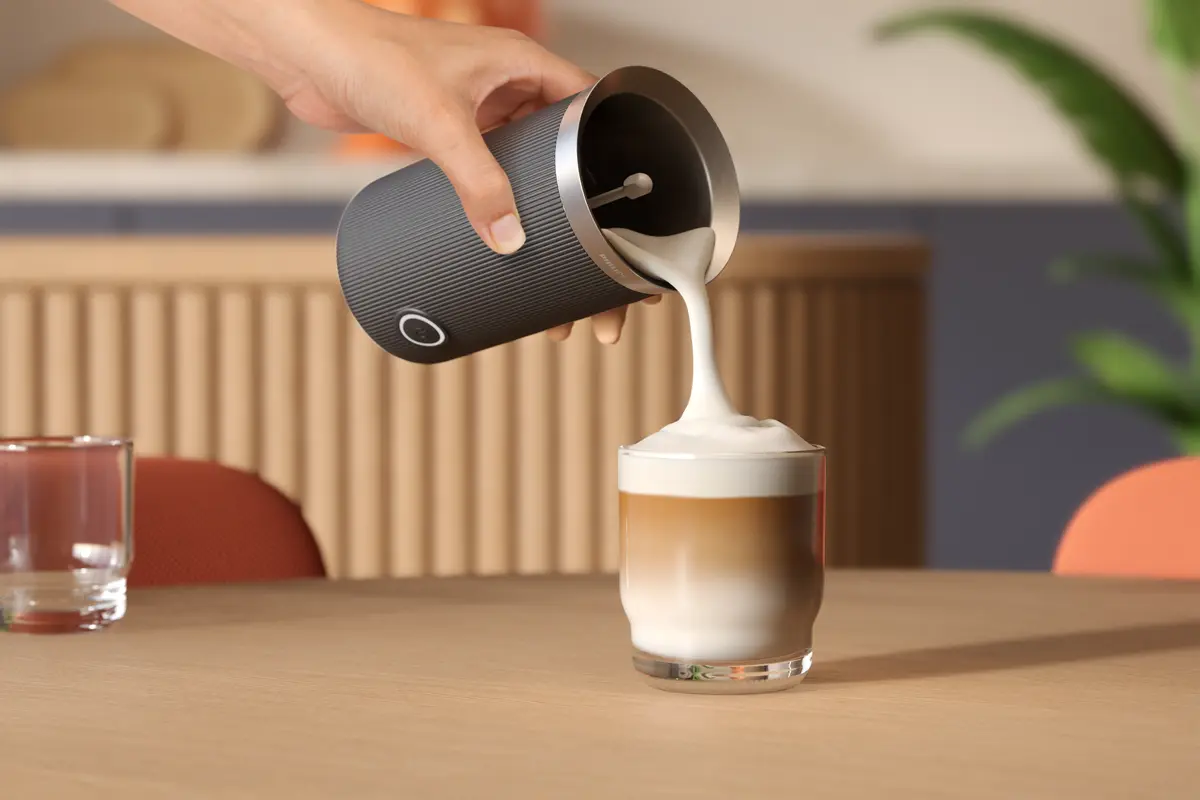 Una mano versa latte schiumoso in una bevanda al caffè a strati. Un montalatte, un bicchiere e un tavolo sono nella scena.