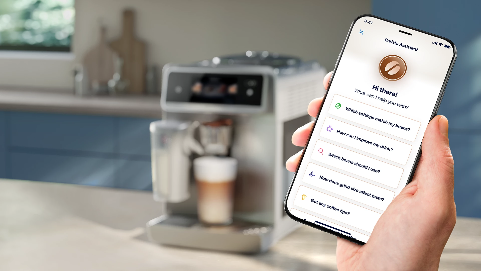 Eine Hand hält ein Telefon mit einer Barista-Assistent-App. Im Hintergrund eine Kaffeemaschine und ein Latte.