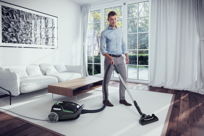Un homme passe l'aspirateur dans un salon. Un canapé, une table basse et une grande fenêtre sont visibles. L'aspirateur est noir et vert.