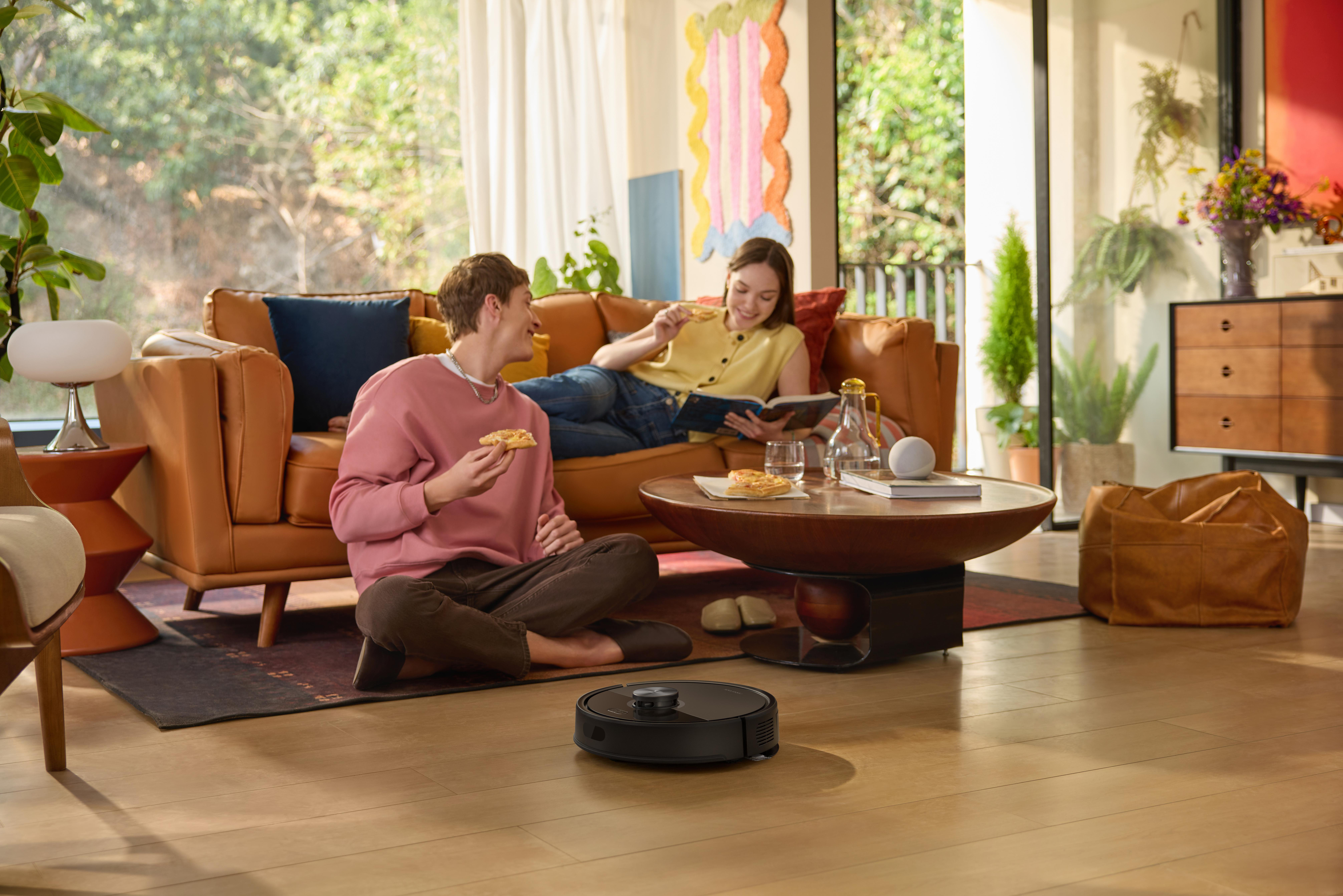 Zwei Personen auf einem Sofa, essen. Ein Roboter-Staubsauger steht auf dem Boden. Ein Couchtisch steht in der Mitte.
