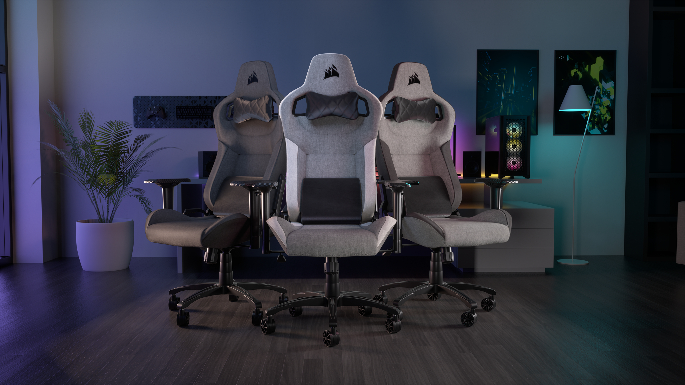 Drei Gaming-Stühle in einem Raum. Einer grau, einer schwarz und einer hellgrau. Eine Pflanze und ein Computer sind ebenfalls im Raum.