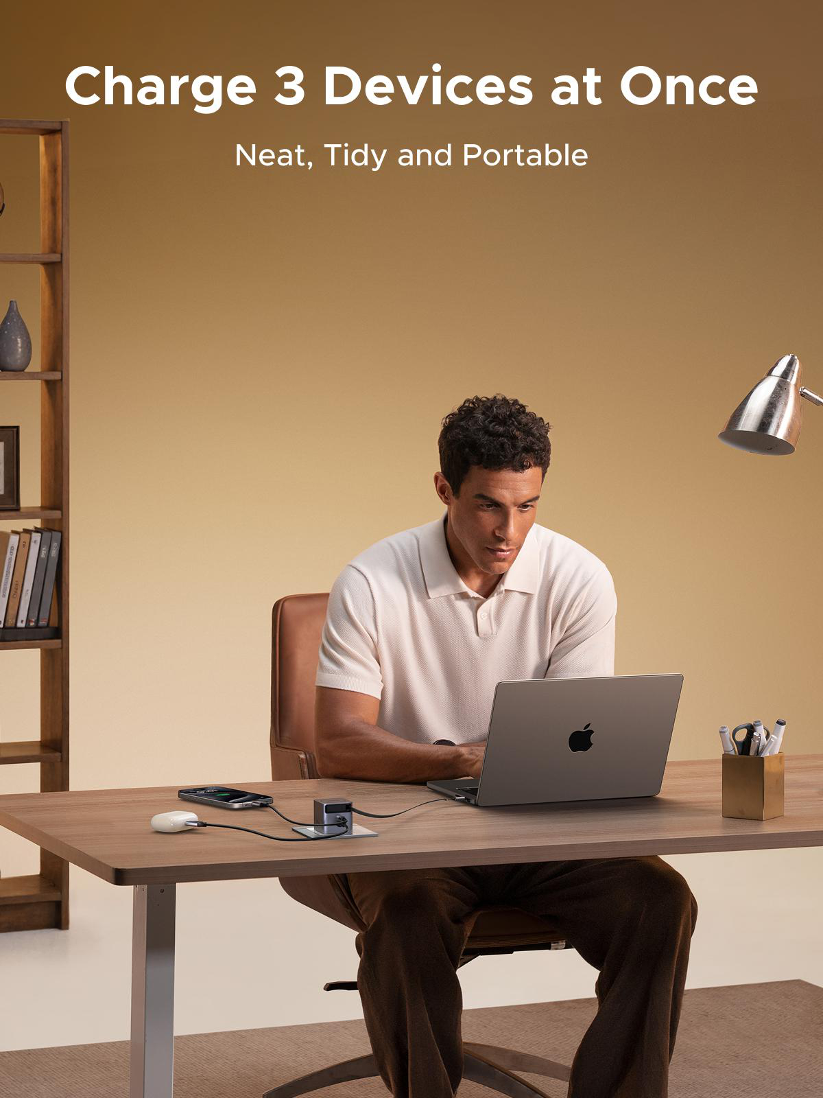Ein Mann sitzt an einem Schreibtisch mit einem Laptop. Auf dem Schreibtisch befinden sich Ladegeräte. Im Hintergrund befinden sich ein Bücherregal und eine Lampe.