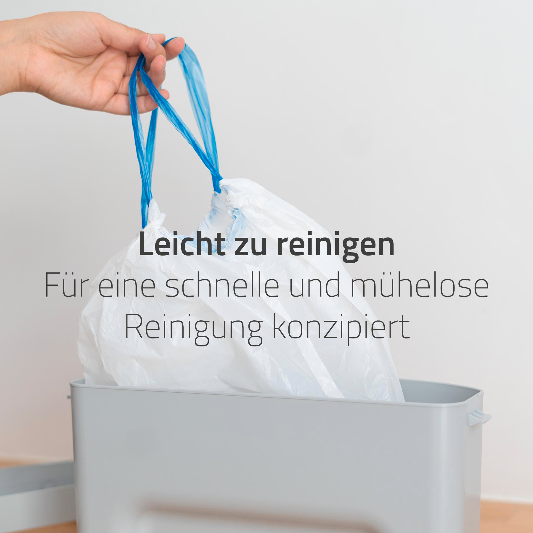 Eine Hand hält einen blaugriffigen Müllsack aus weißem Kunststoff. Der Beutel befindet sich über einer grauen Tonne.