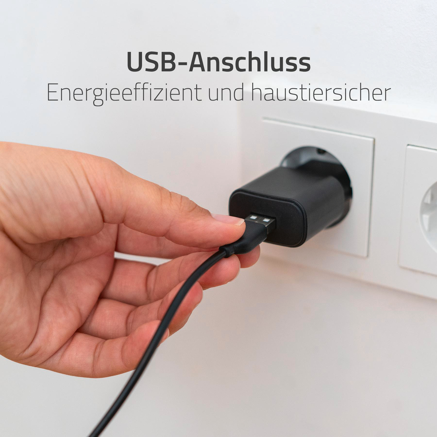 Eine Hand, die ein USB-Kabel in ein schwarzes Ladegerät in einer Steckdose steckt. Text: 'USB-Anschluss'.