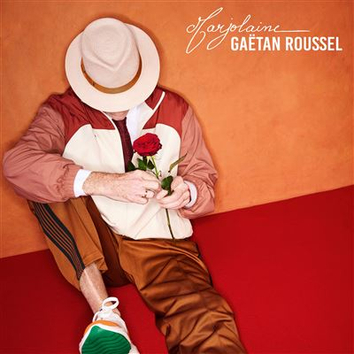Homme avec chapeau tenant une rose rouge, assis sur une surface rouge sur fond orange. Texte: 'Marjolaine Gaëtan Roussel'.