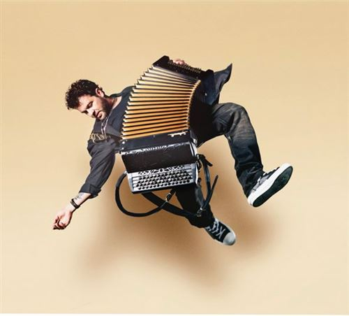 Un homme en l'air joue de l'accordéon. Il porte une veste noire, un jean foncé et des baskets. Le fond est beige clair.