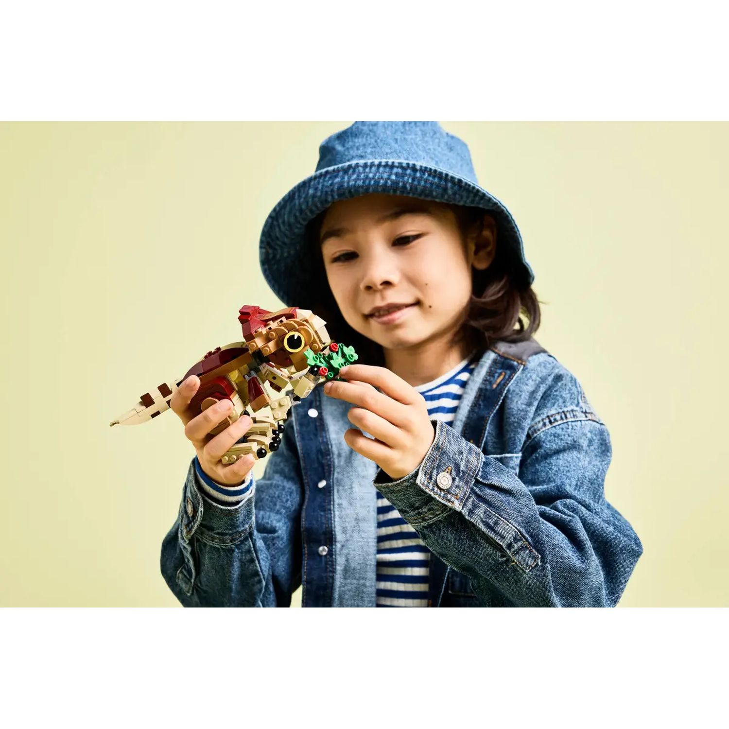 A child in a denim jacket and hat holds a Lego dinosaur, smiling. The dinosaur is brown, beige, and red. Background is light yellow.