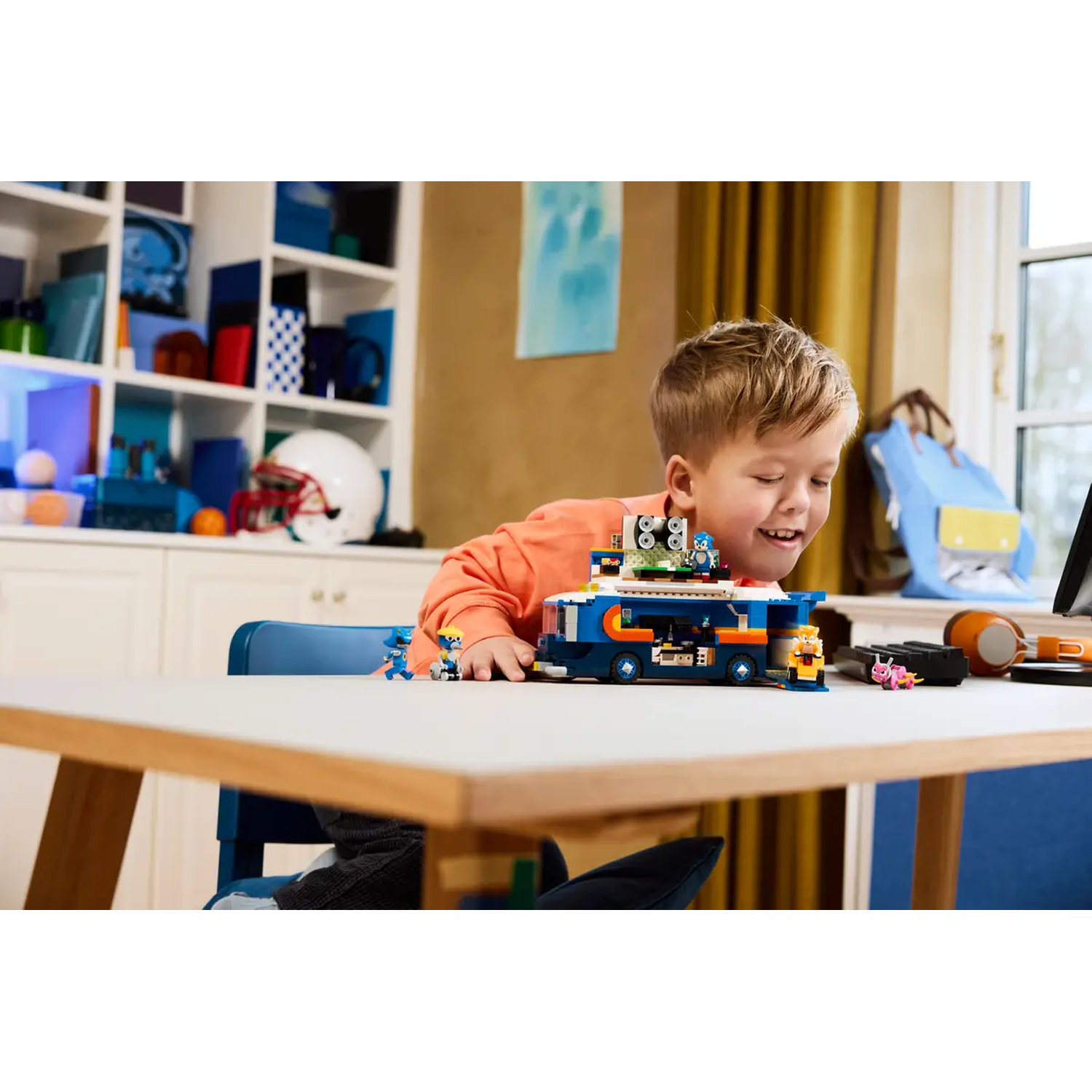 Een jongen lacht terwijl hij speelt met een blauw-witte speelgoedbus. Er staan verschillende speelgoedfiguren en een laptop op tafel.