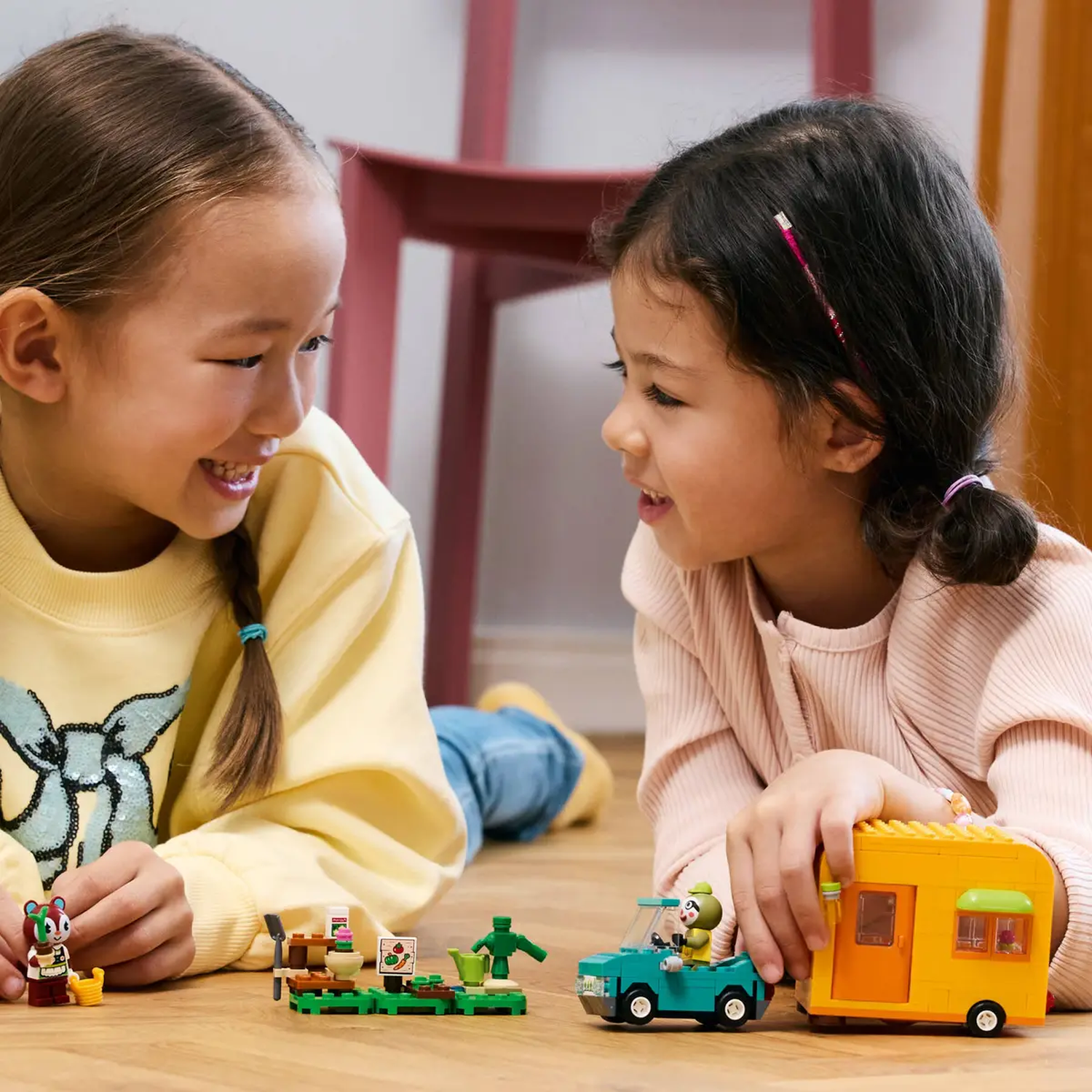 Twee meisjes lachen naar elkaar, spelend met Lego-speelgoed van een auto, een caravan en een tuin. De scène is binnen.