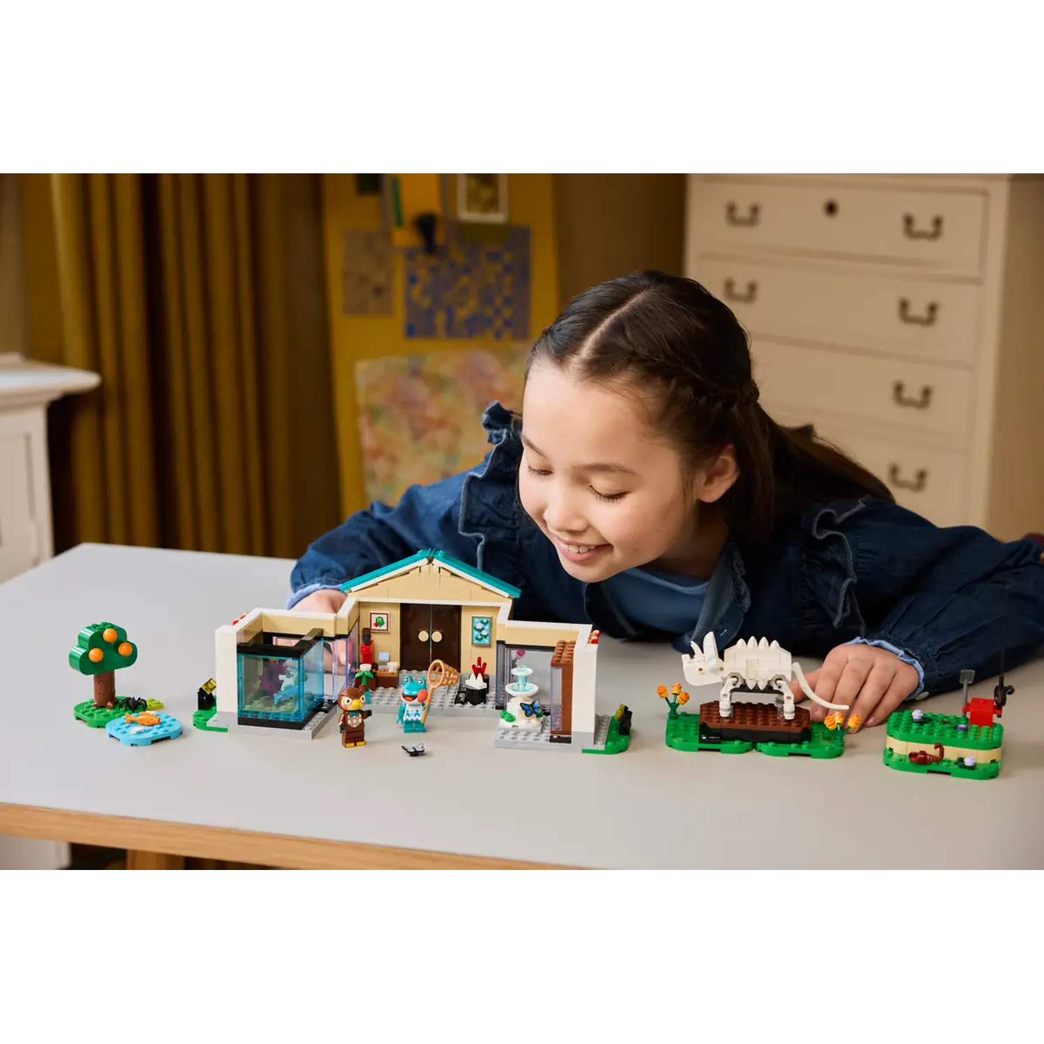 Een meisje bekijkt een Lego-set. De set bevat een huis, figuren en een dinosaurus. Ze glimlacht, gebogen over de set op een tafel.