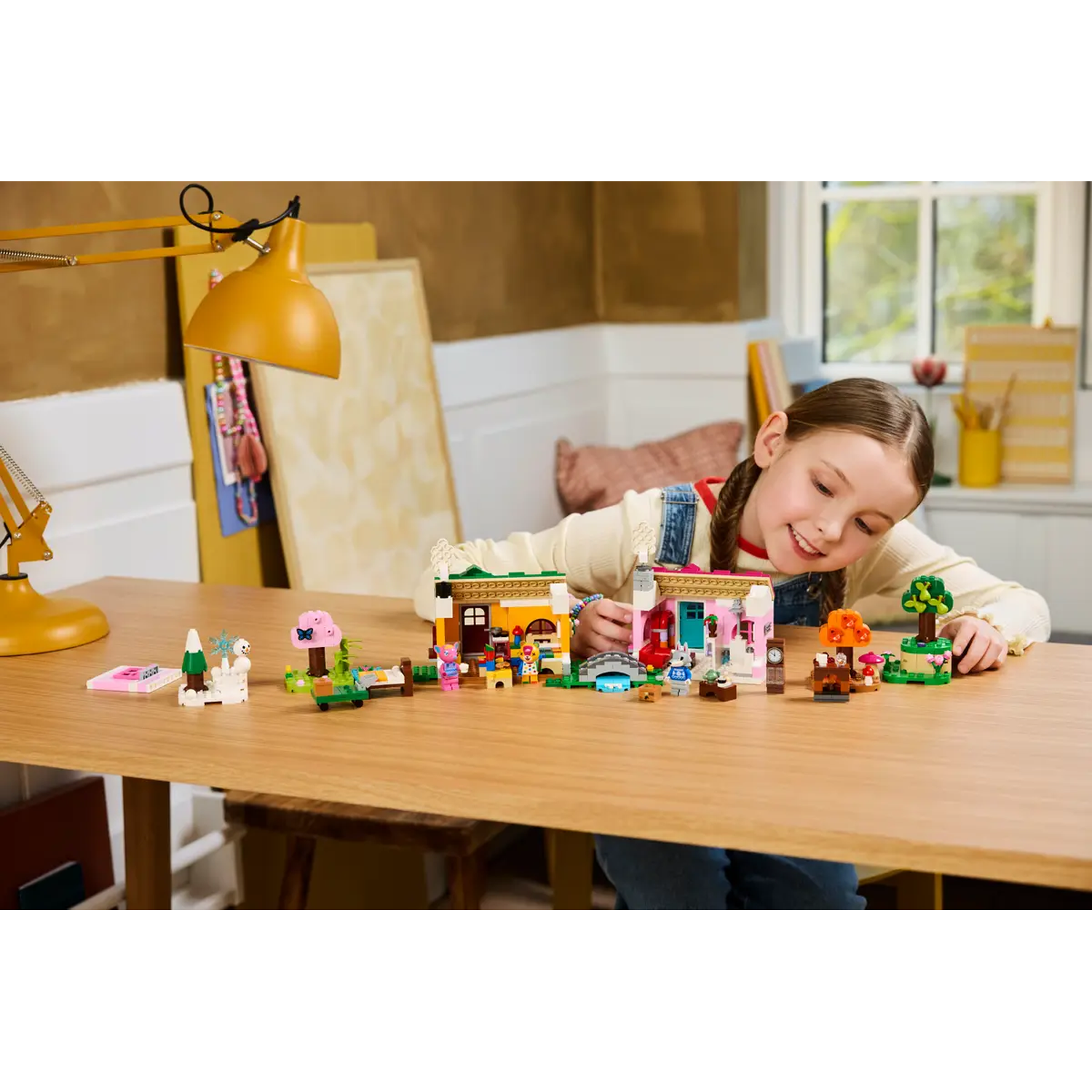 Een meisje glimlacht naar een Lego-stad op een tafel. De stad heeft huizen, bomen en figuren. Een gele lamp staat links.
