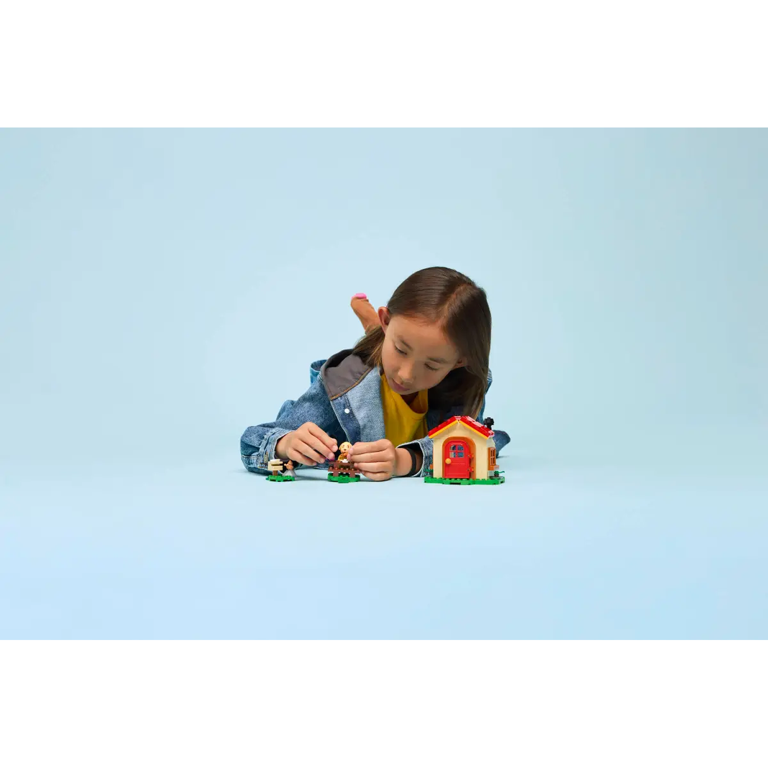 A girl lies on the floor playing with toy blocks. There is a house and two figures made of blocks. The background is light blue.