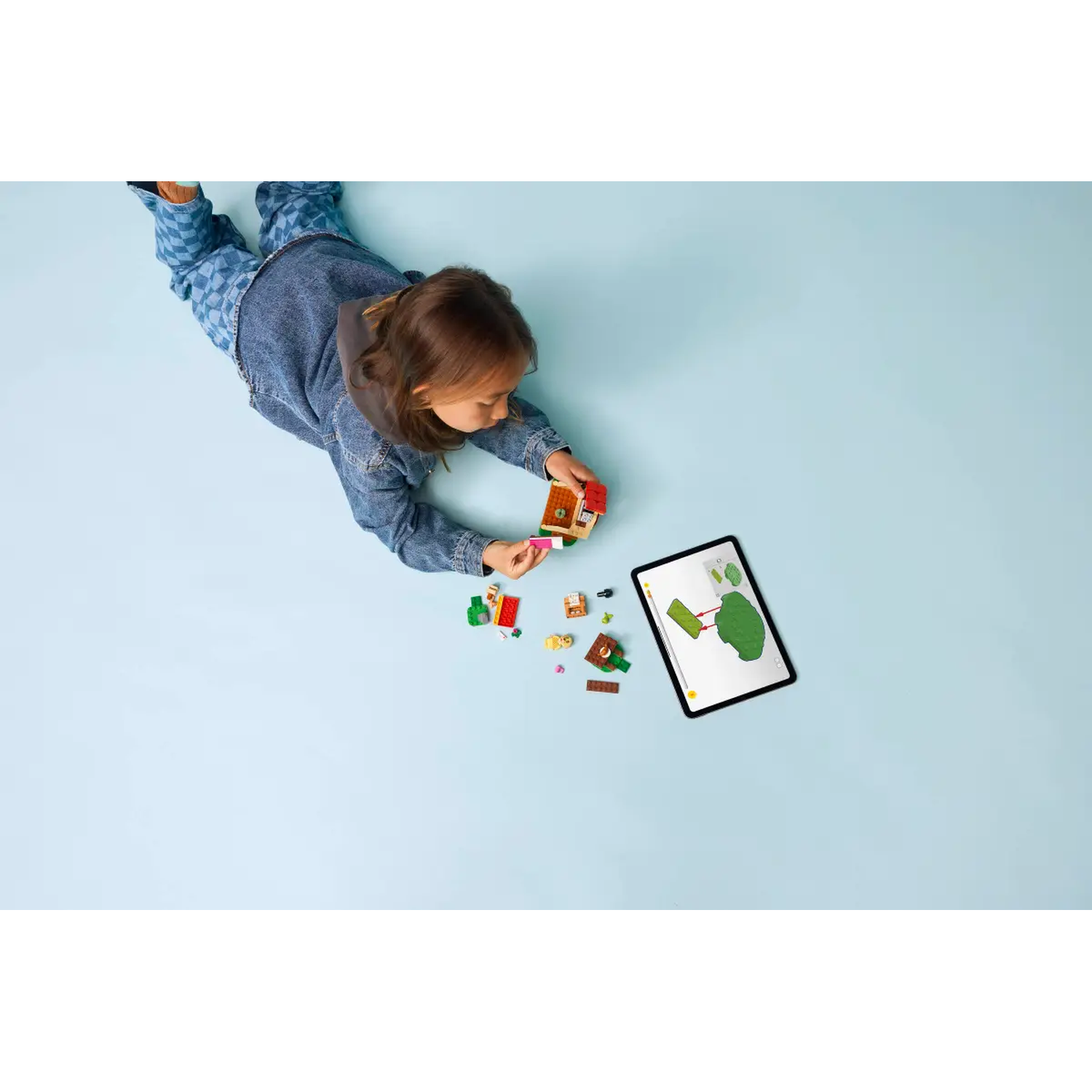 A child in denim plays with LEGO bricks, a tablet shows instructions. Bricks and a small house are scattered on a blue surface.
