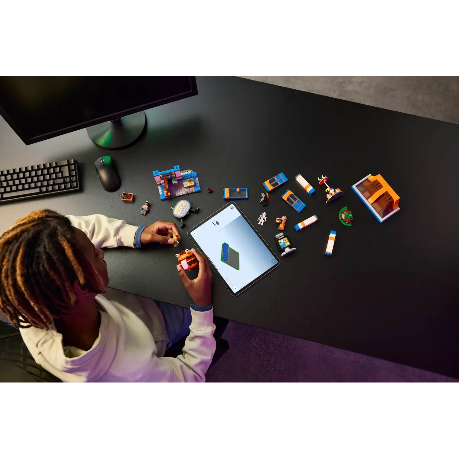 Een persoon speelt met LEGO's op een zwart bureau. Een tablet, toetsenbord en monitor staan ook op het bureau.