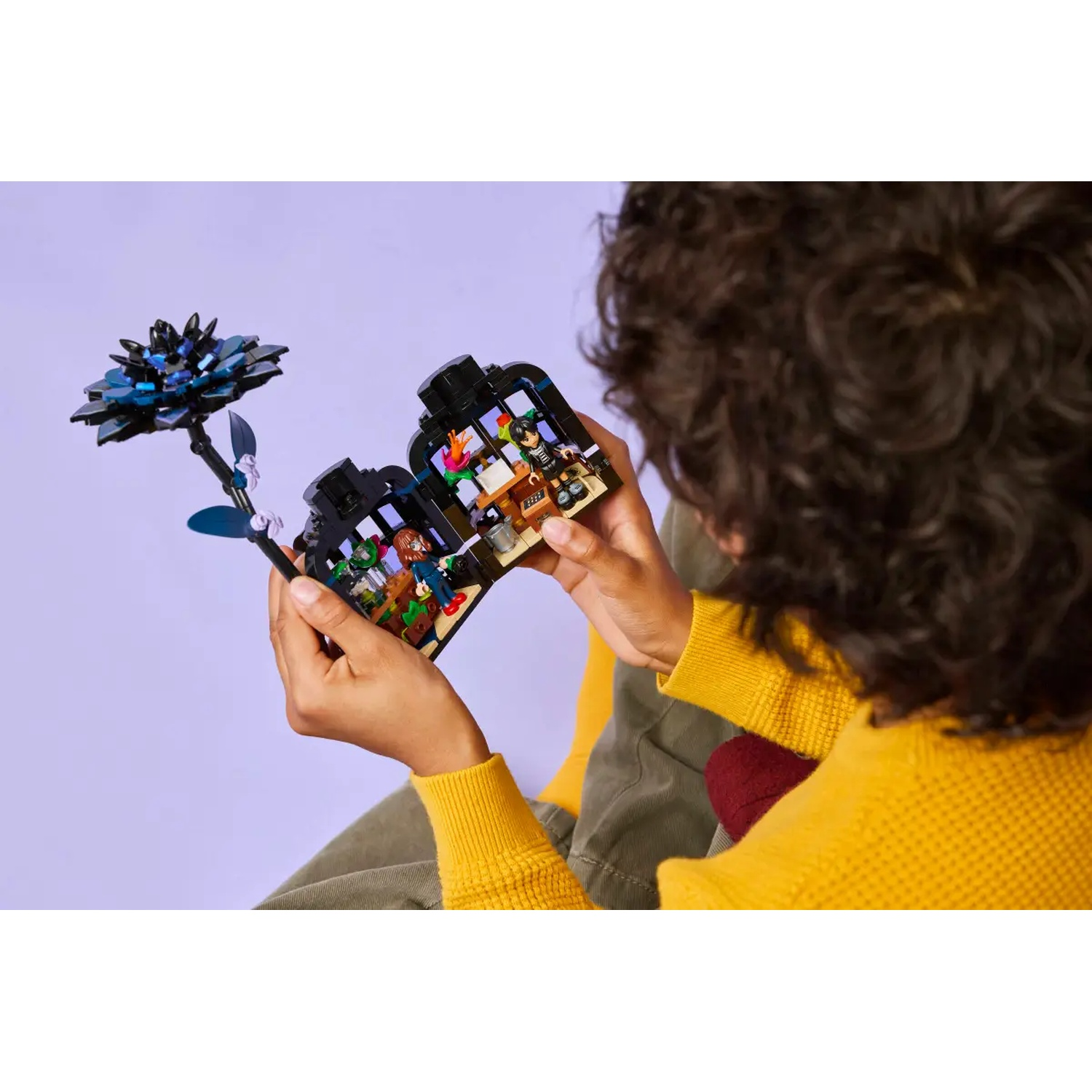 Une personne tient un set Lego avec une fleur et une pièce miniature. La personne porte un pull jaune. Le fond est violet.