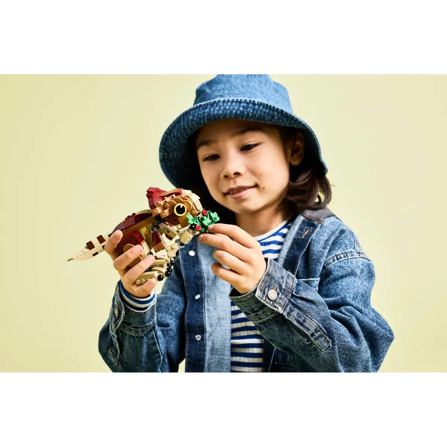 A young person in a denim jacket and hat holds a Lego dinosaur, smiling. Green background.