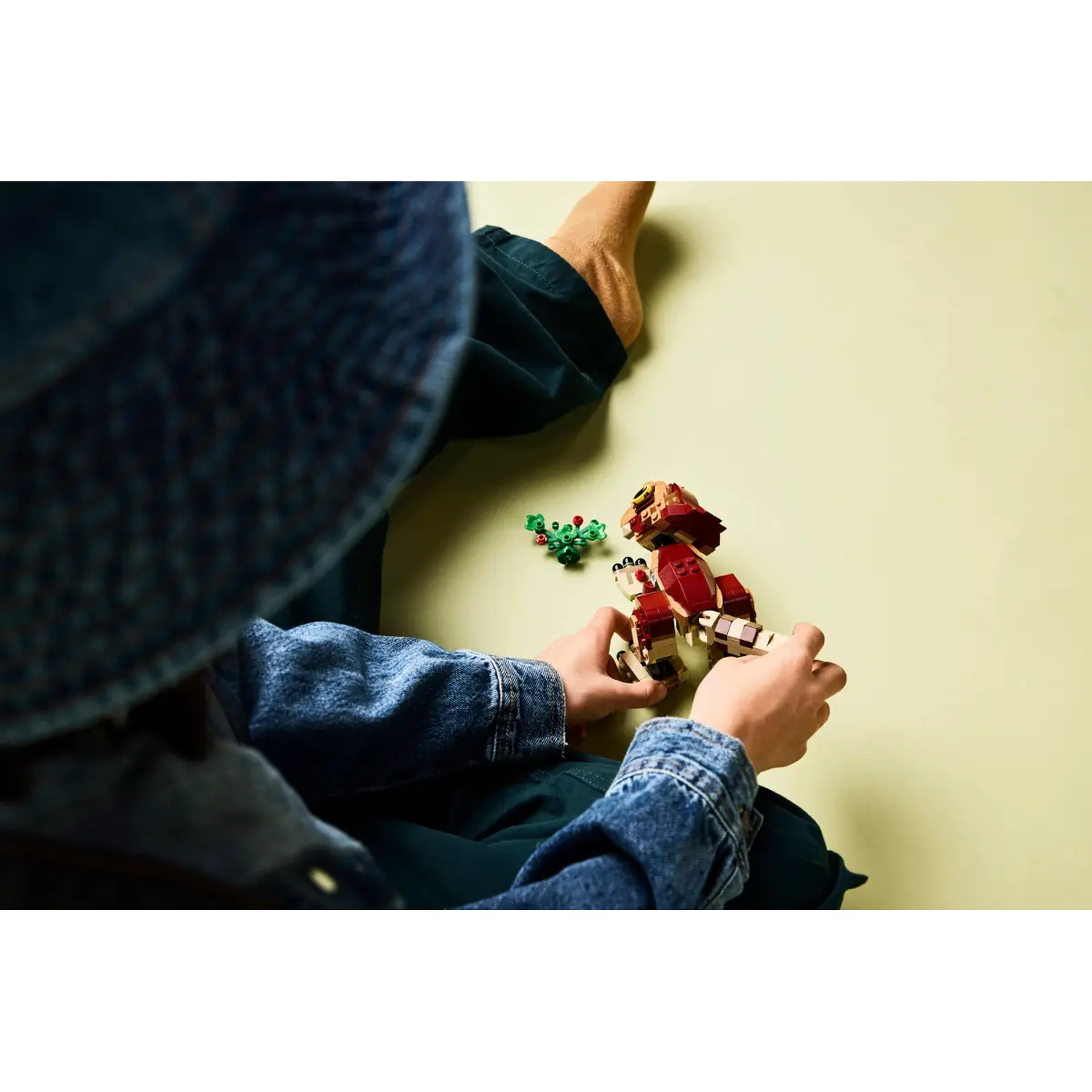 A person in denim jacket and hat plays with a red and beige Lego robot, next to a small green Lego plant. Green background.