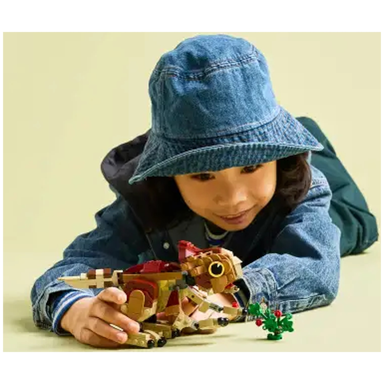 A child in a denim jacket and hat looks at a Lego dinosaur toy. A small Lego plant is beside the toy.
