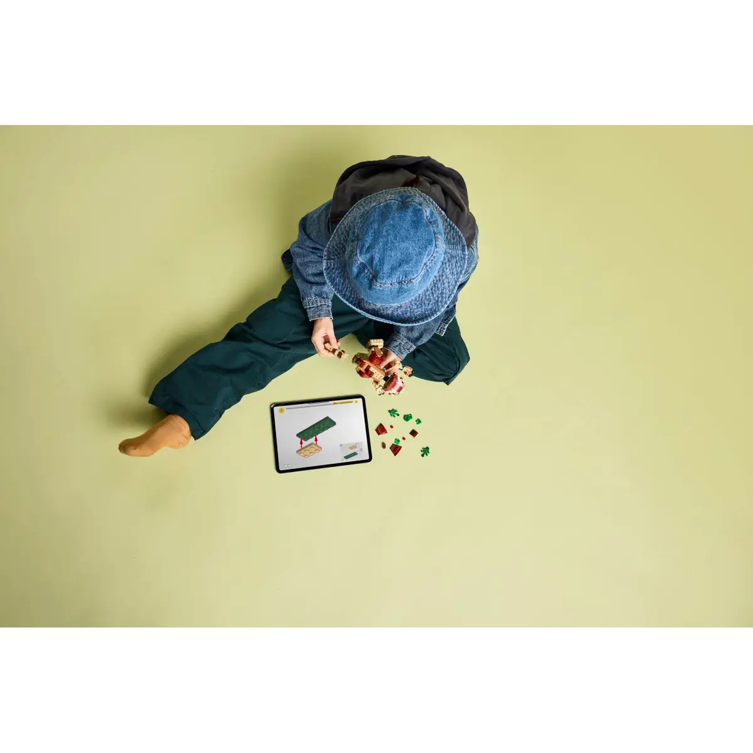 A child in a denim hat and green pants is playing with colorful blocks. A tablet shows instructions. Green background.