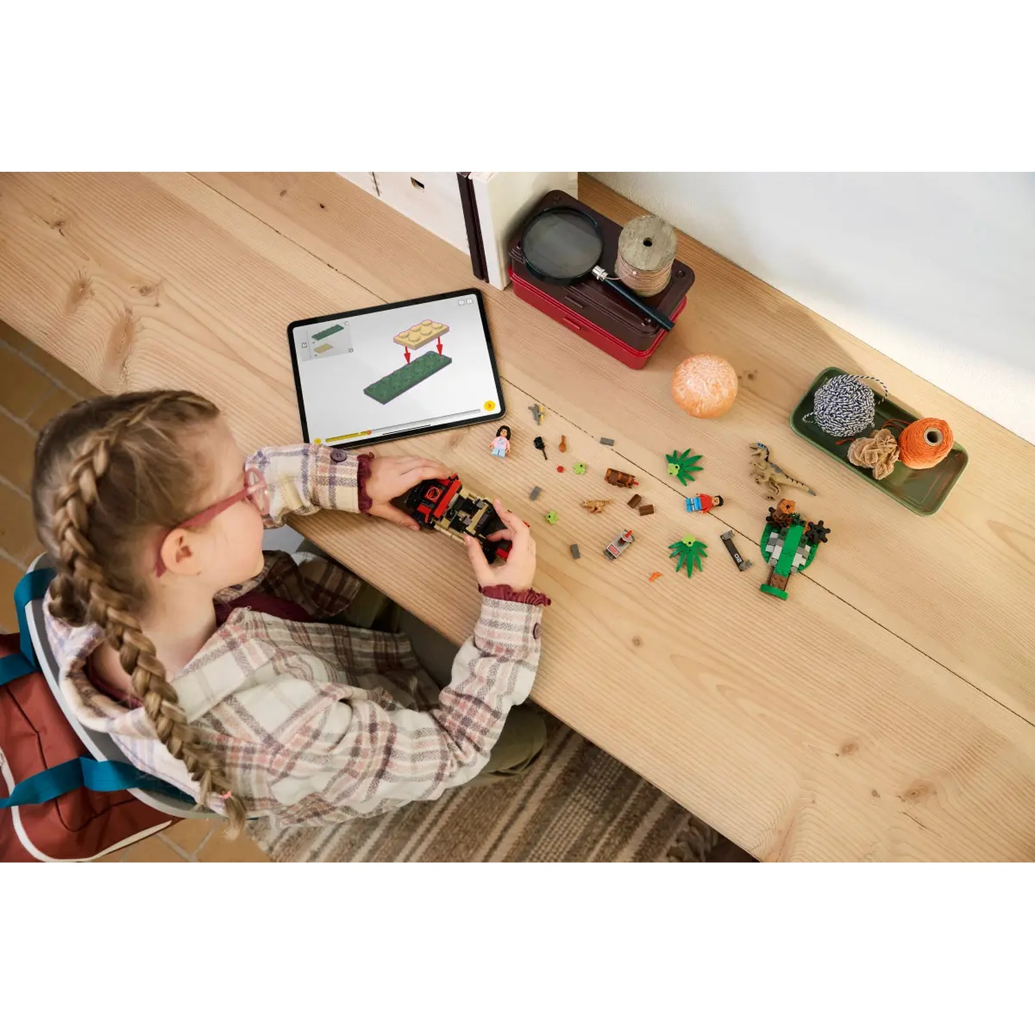 A girl with braids looks at a tablet showing building instructions. Lego pieces and other objects are on the wooden table.