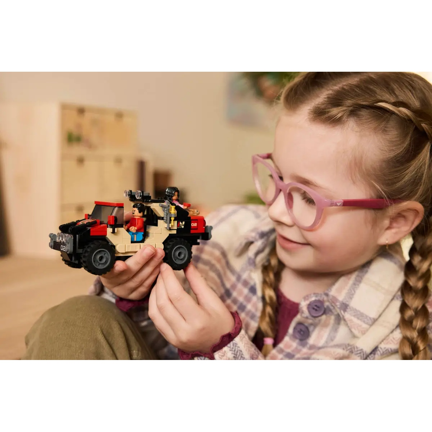 A girl with glasses holds a Lego car. The car is black, red and beige with Lego figures inside. The girl smiles.