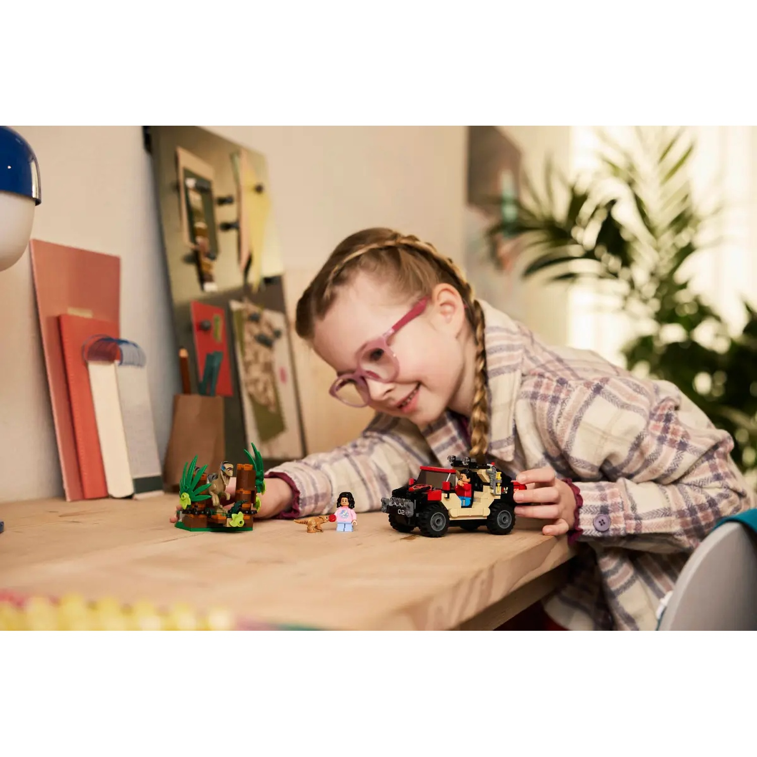 A girl with glasses plays with Lego. There's a car, a dinosaur, and a minifigure on a wooden desk. Background includes books.