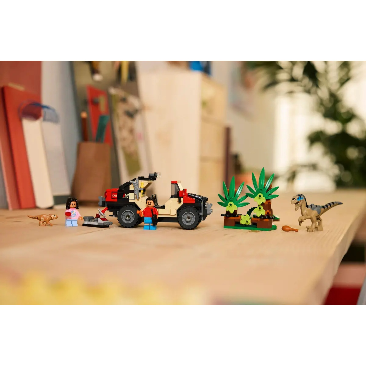 Lego Jurassic Park set on a table. Includes figures, a jeep, dinosaurs, and plants. The background shows blurred books and furniture.