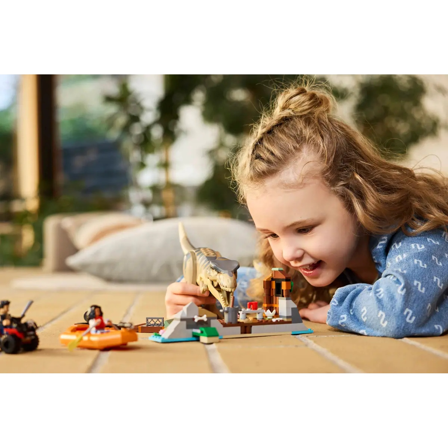 A young girl smiles while playing with a dinosaur Lego set. The set includes figures, a boat, a car, and building blocks.
