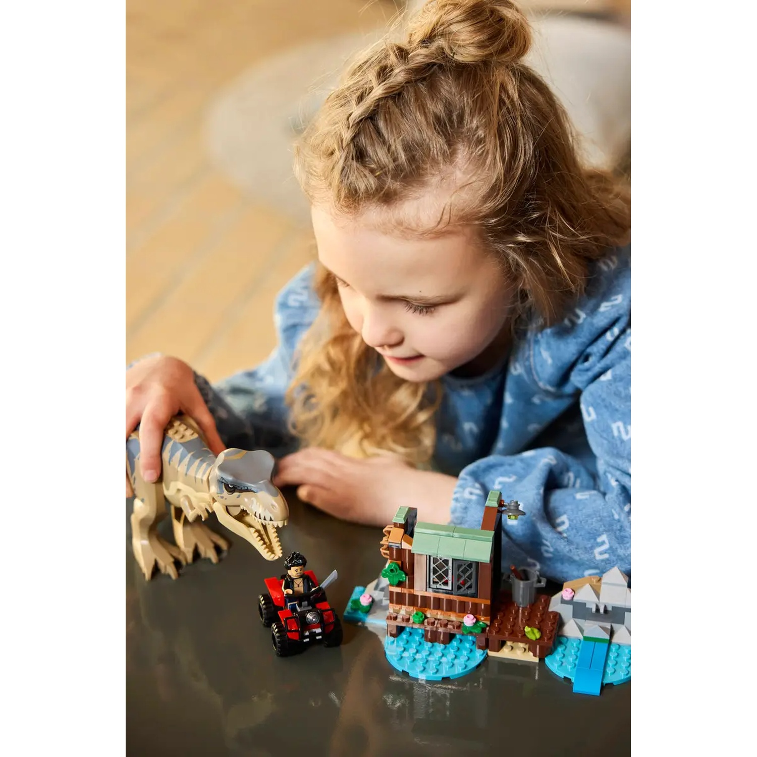 A girl looks at a Lego set with a T-Rex, a red ATV, a small house, and a water feature.