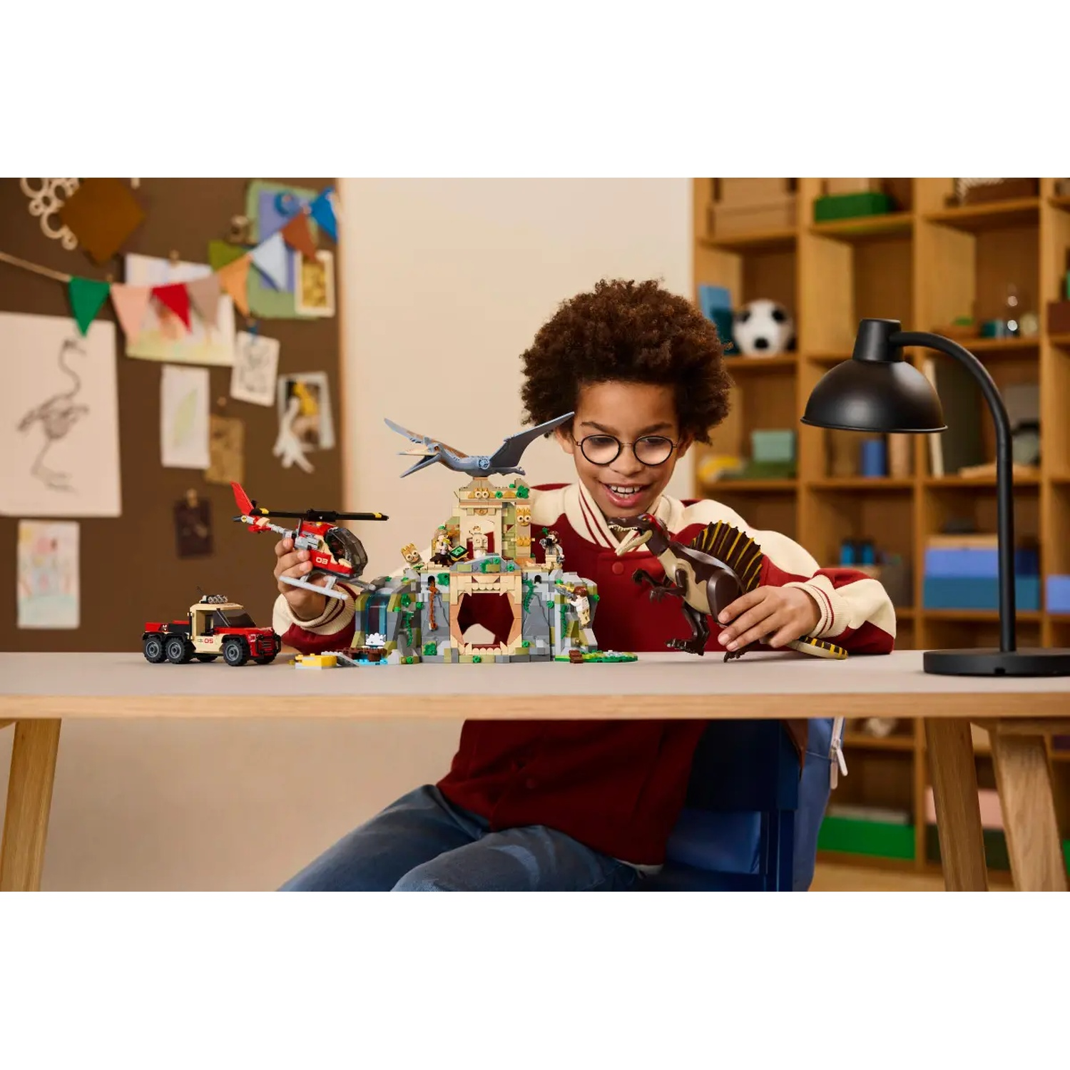 A boy with glasses plays with dinosaur toys. A red helicopter, a truck, and a Lego structure are on the table. A lamp is on the right.