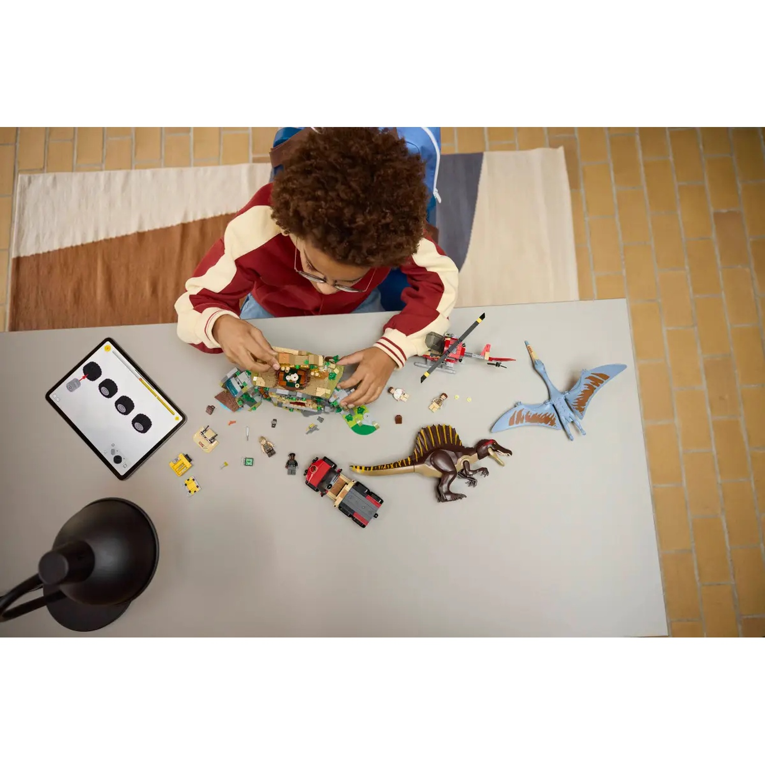 A child is playing with dinosaur and helicopter toys on a table. There's a tablet, a lamp, and a rug visible.