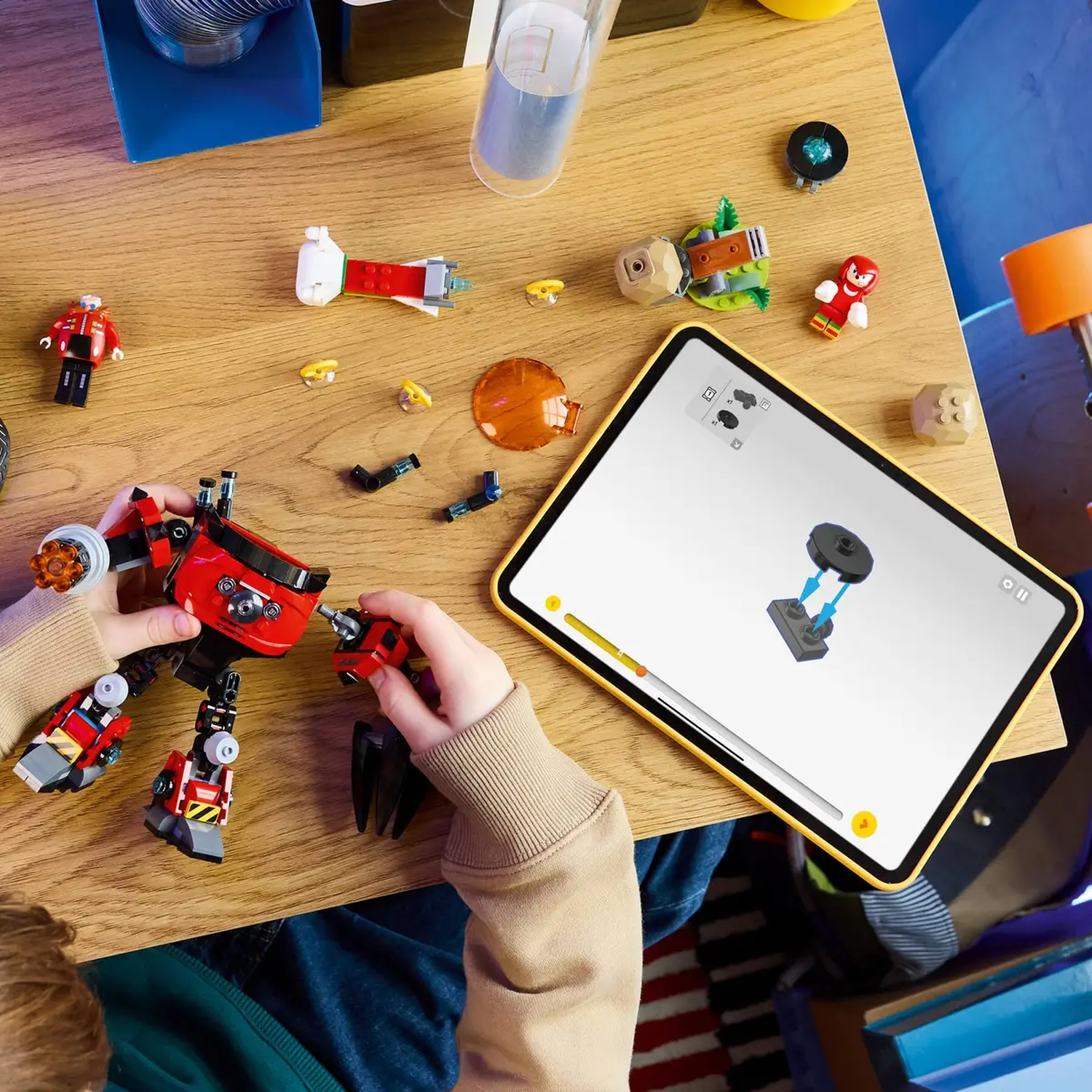 Un enfant assemble un robot Lego rouge et noir. Les instructions sont sur une tablette. Des pièces et figurines Lego sont éparpillées sur la table en bois.