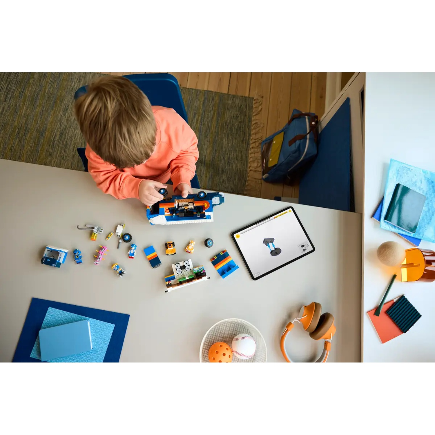 Een kind assembleert een speelgoedauto op een tafel, omringd door diverse speelgoedonderdelen, een tablet en een koptelefoon.