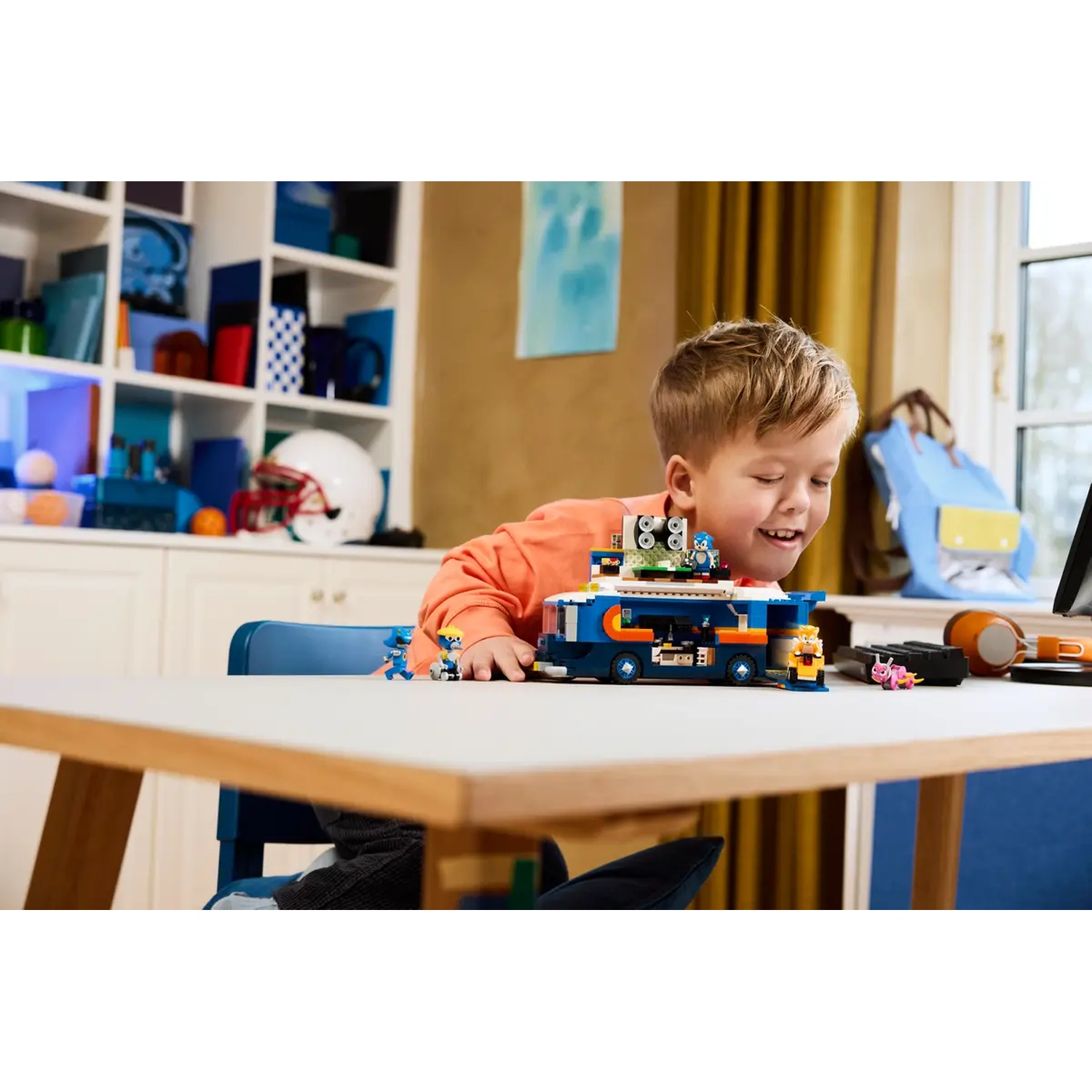 Een jongen lacht naar een Lego-set, met een blauw voertuig en Lego-figuren. Een tafel, stoel en achtergrond schappen zijn zichtbaar.