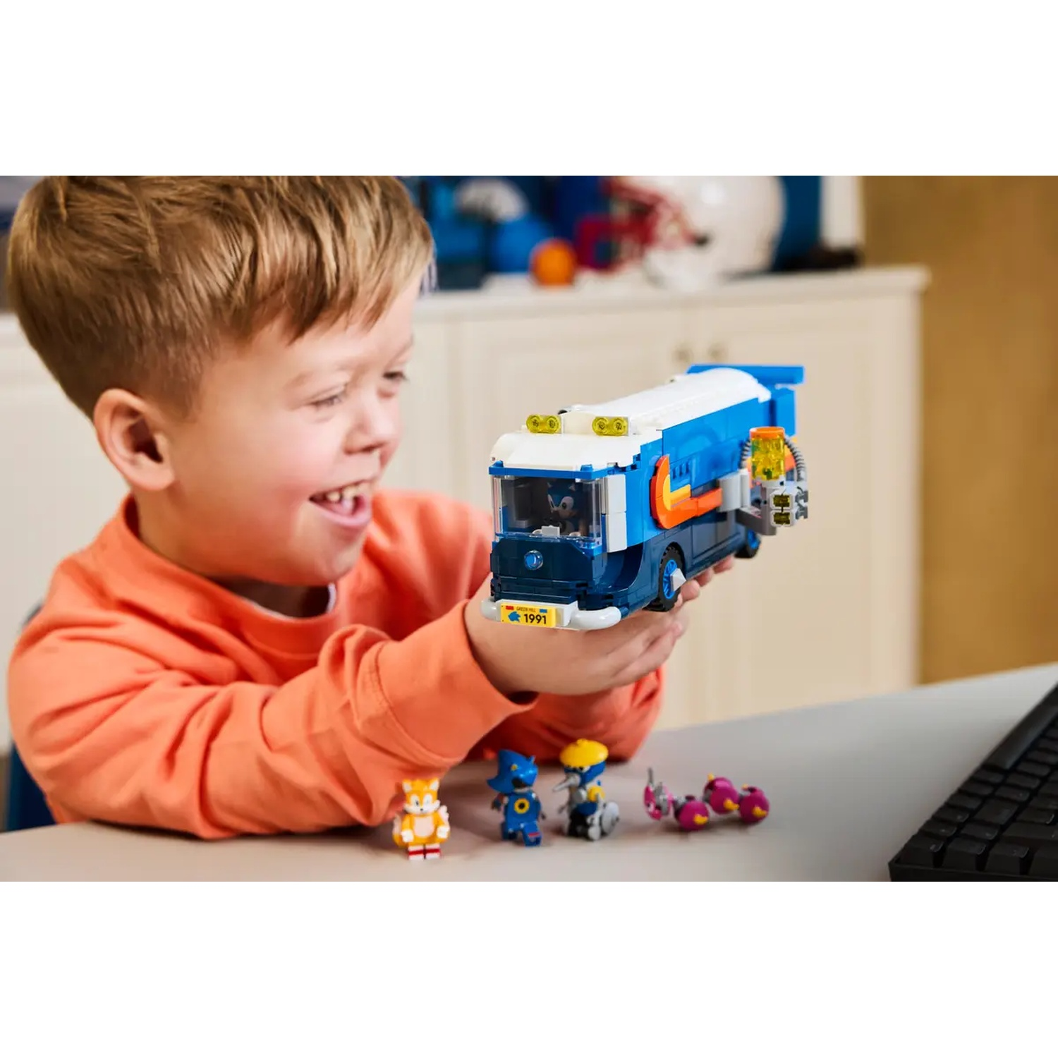 Een lachende jongen houdt een blauwe en oranje speelgoedbus vast. Kleine speelgoedfiguren staan op tafel. Een toetsenbord staat op de achtergrond.