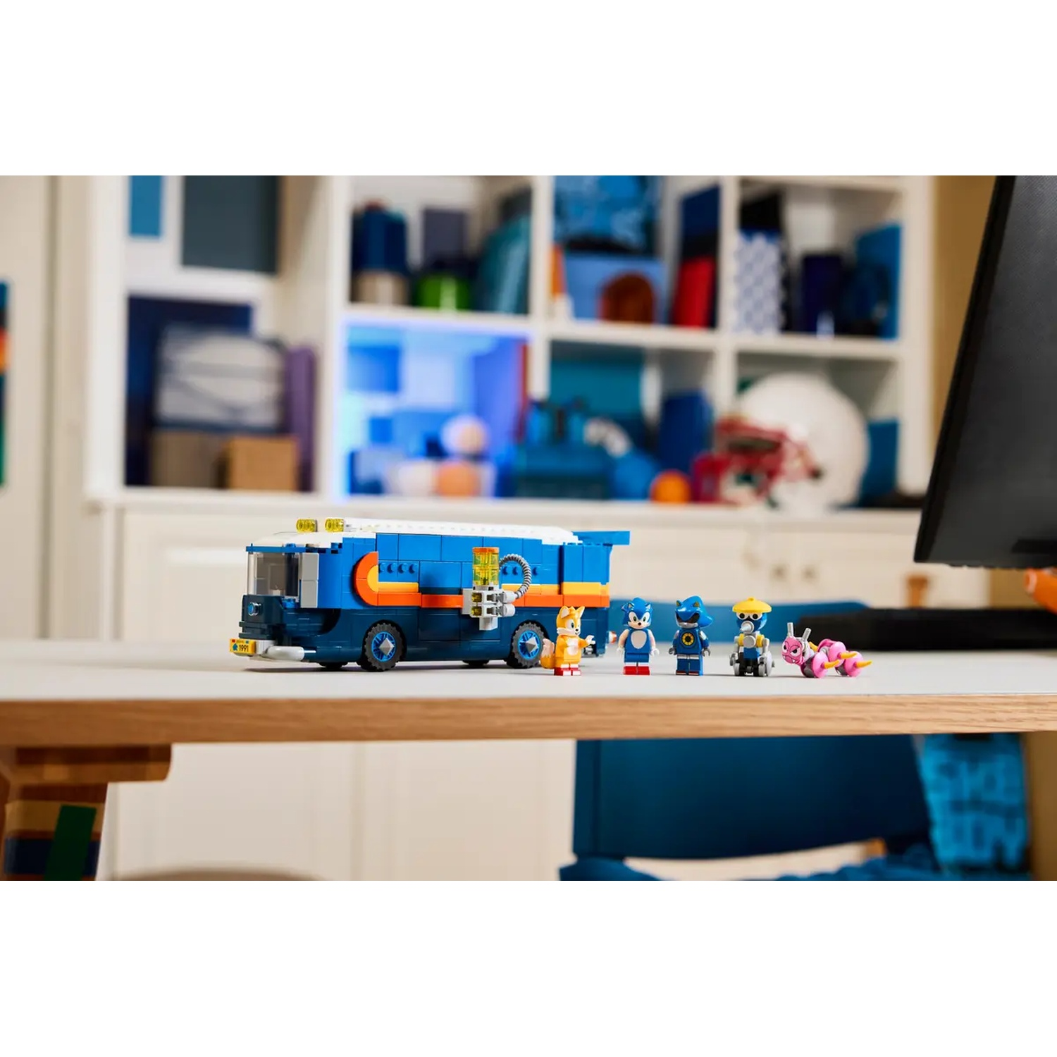 Een blauwe Lego-bus en Sonic-personages staan op een tafel. De achtergrond omvat een witte plank en een zwart computerscherm.