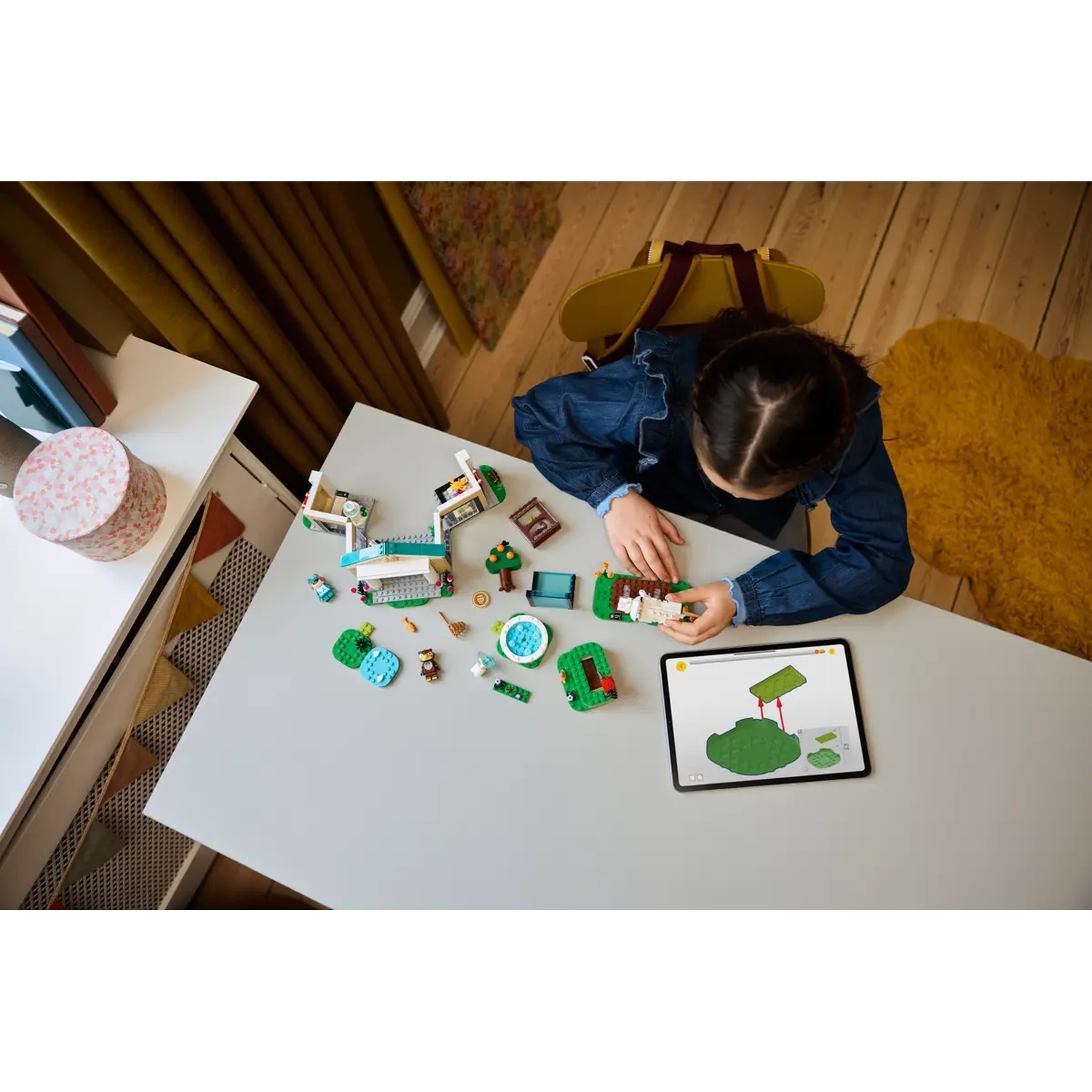 Une fille assemble un jeu Lego sur une table. Des pièces Lego et une tablette affichant des instructions sont visibles. Tapis et rideaux jaunes en arrière-plan.
