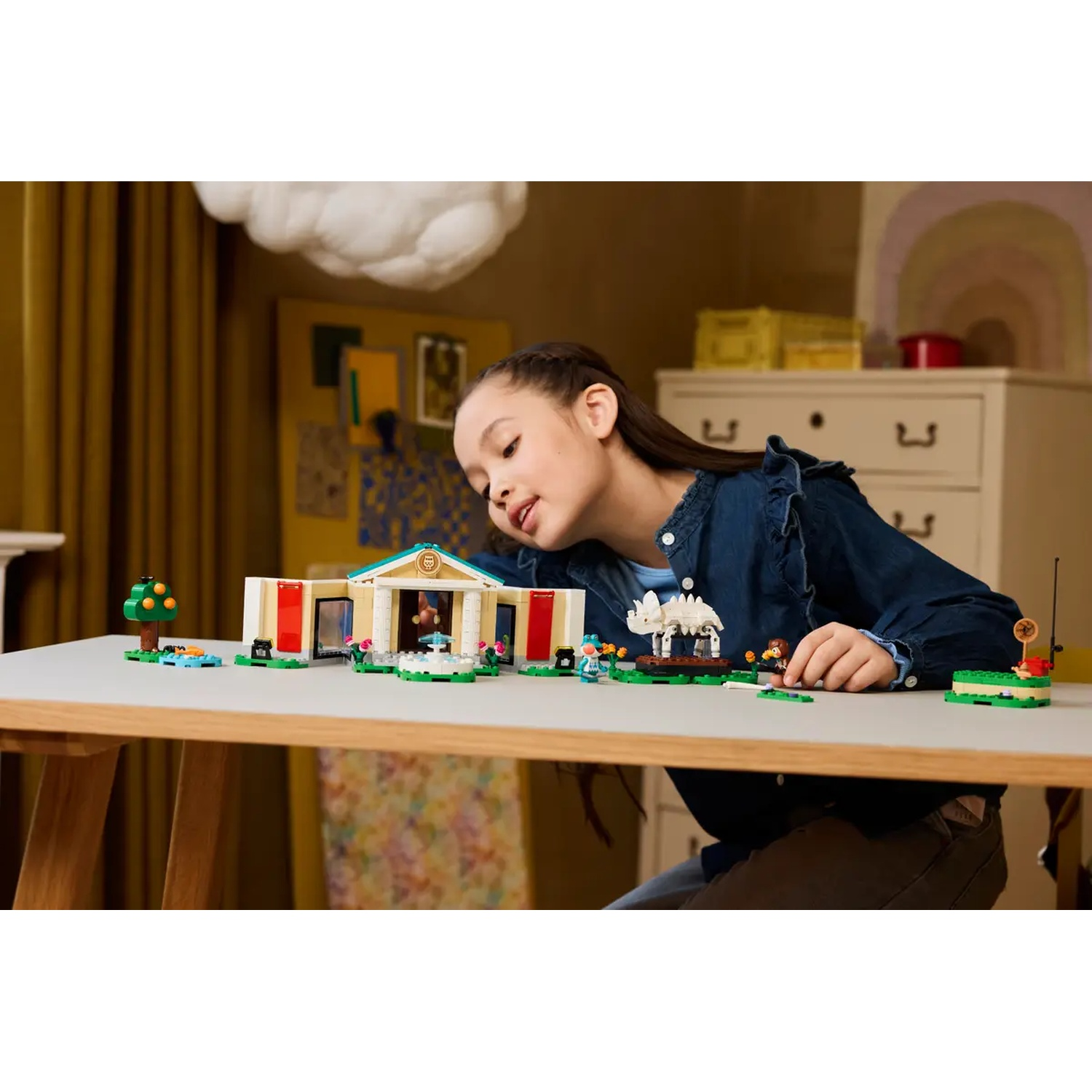 Une jeune fille se penche sur un jeu de Lego sur une table. L'ensemble comprend un bâtiment, un arbre et de petites figurines. Le fond a une déco.