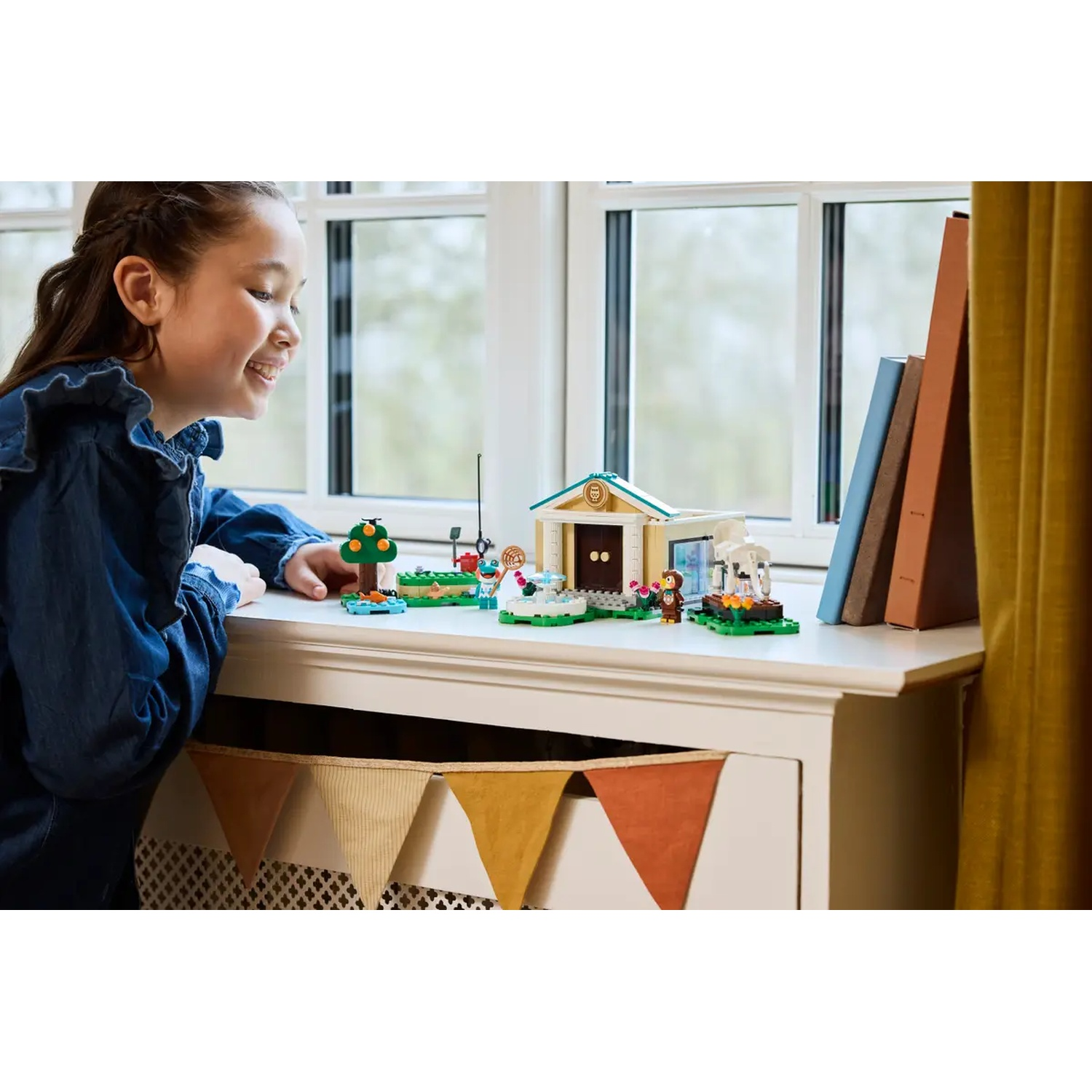 Une fille regarde un set Lego avec un bâtiment, un arbre et des figurines. Livres et fenêtre en arrière-plan. Une bannière est suspendue en dessous.
