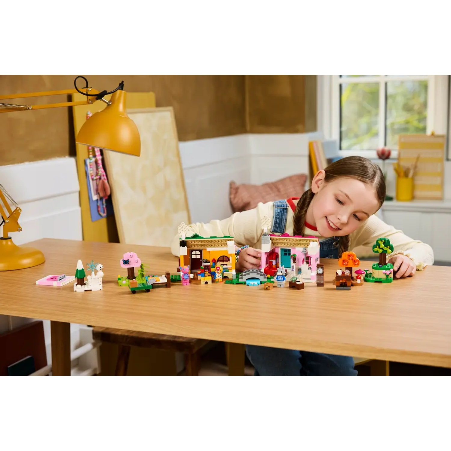 Une fille sourit en jouant avec des ensembles LEGO Friends sur une table en bois. Bâtiments, arbres et petites figurines sont disposés.