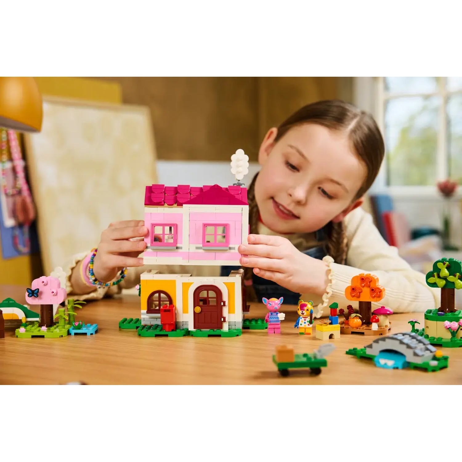 Une fille joue avec des LEGO. Elle construit une maison rose avec une base jaune. Divers personnages et paysages LEGO sont placés.