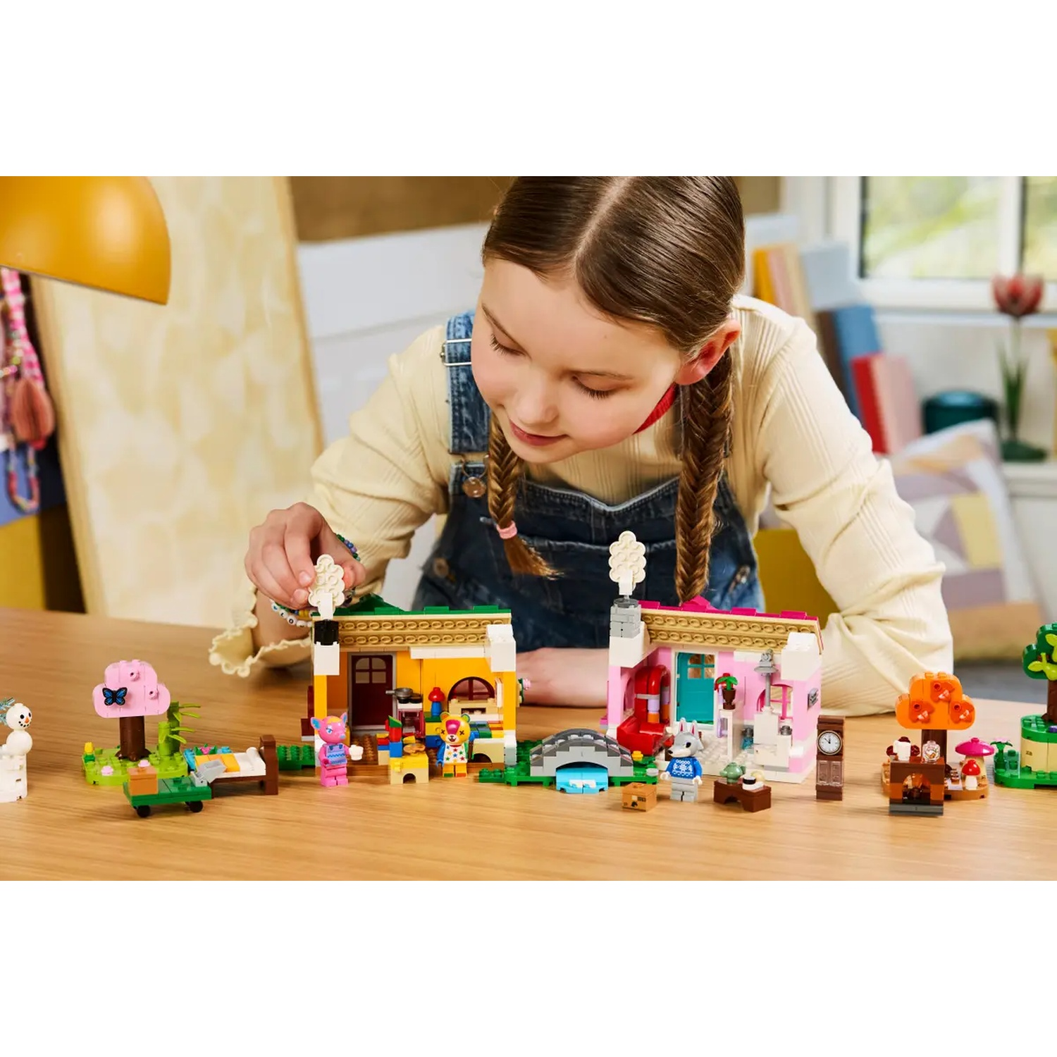 Une fille joue avec des blocs Lego. Elle construit des maisons et un paysage avec diverses figurines et accessoires Lego sur une table en bois.