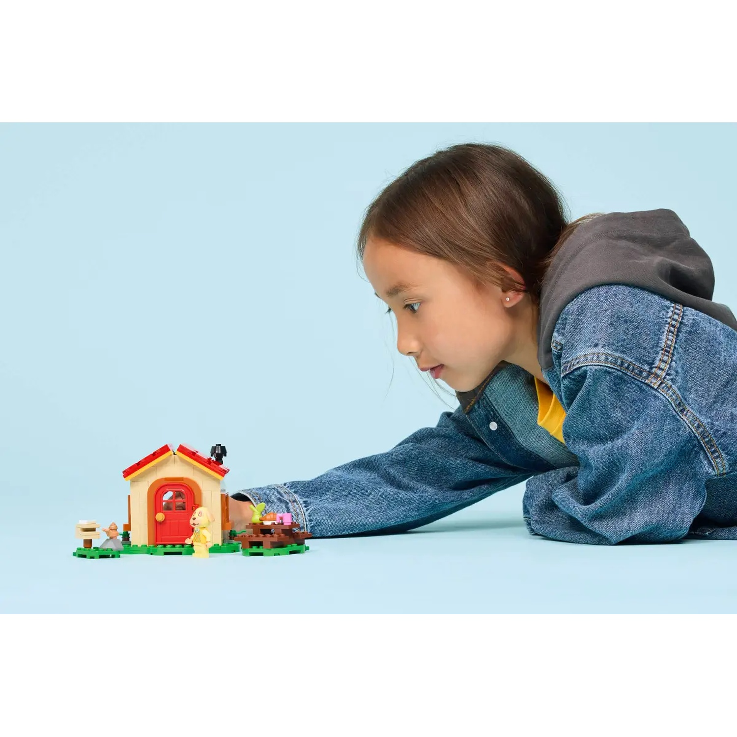 Une fille regarde une maison Lego. La maison est beige et rouge, avec une petite table et une figurine devant. Fond bleu.