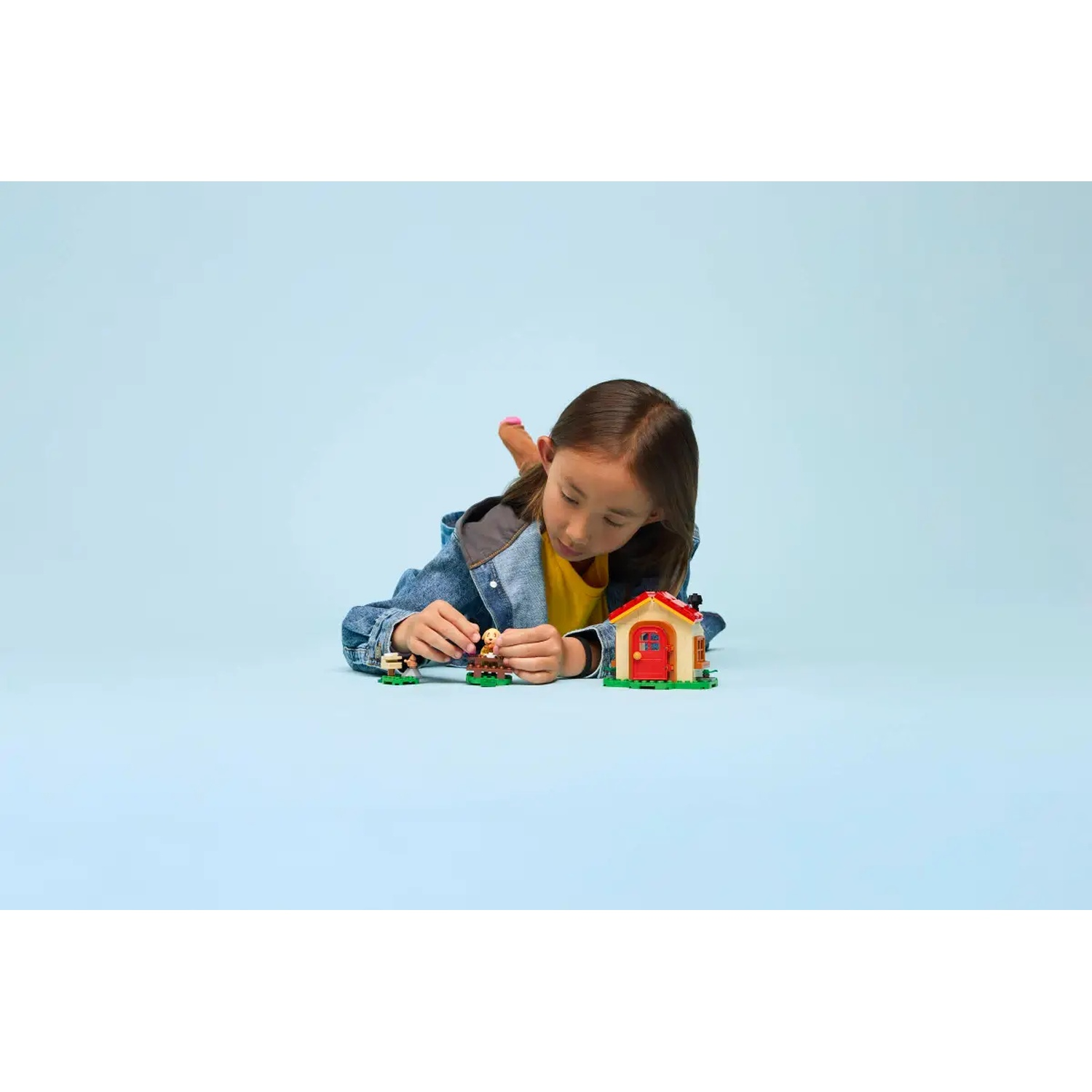 Une fille est allongée sur le sol et joue avec une maison de jouet et des figurines. La maison a un toit rouge et une porte. Fond bleu.