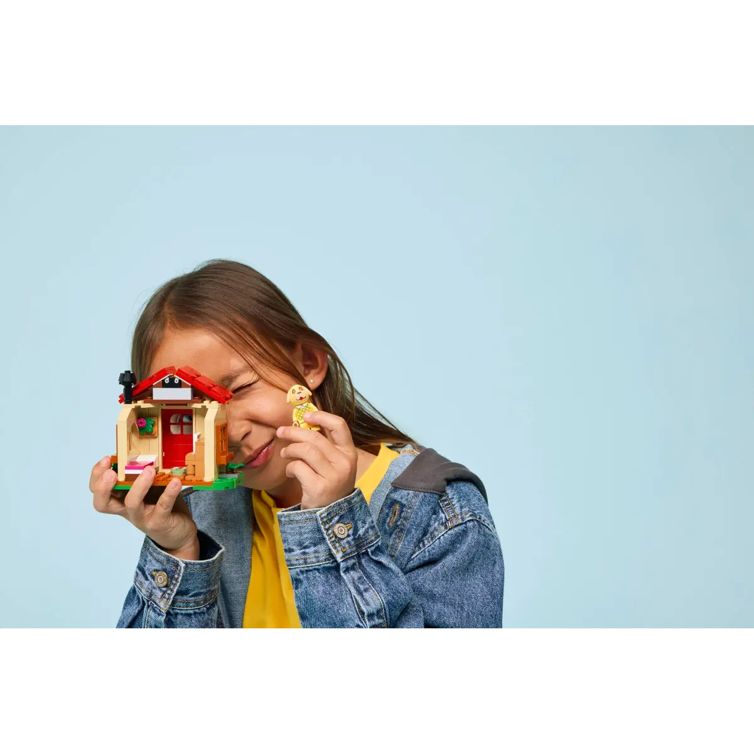 Une fille sourit, tenant une maison Lego et une petite figurine. Elle porte une chemise jaune et une veste en jean, sur fond bleu.