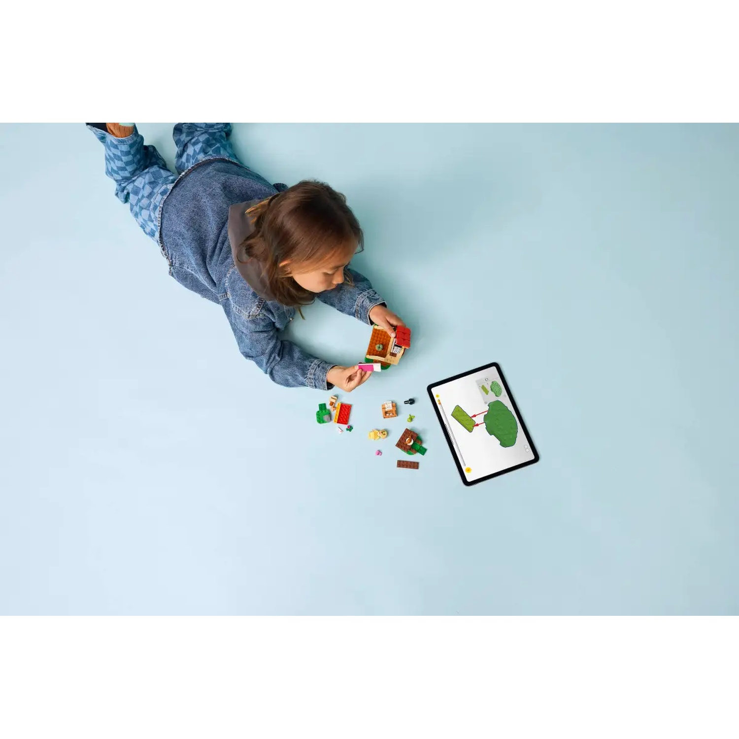 Un enfant en jean joue avec des LEGO, une tablette affichant une image d'arbre est à proximité sur une surface bleu clair.