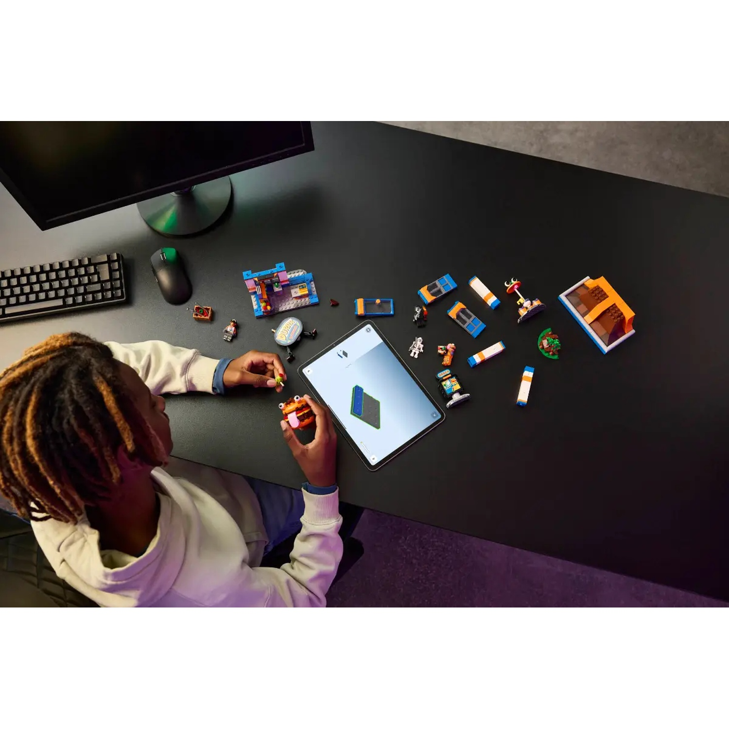 Une personne joue avec des LEGOs sur un bureau noir. Un moniteur, un clavier et une souris sont visibles. Une tablette est utilisée.