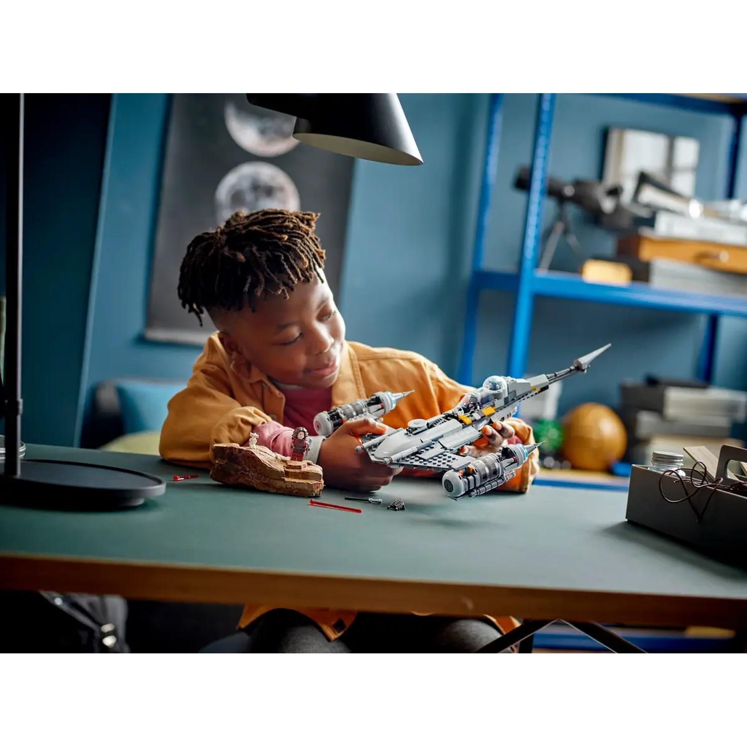 A young boy with dreadlocks is assembling a Star Wars Lego spaceship on a green desk. Background shows a shelf and wall decor.