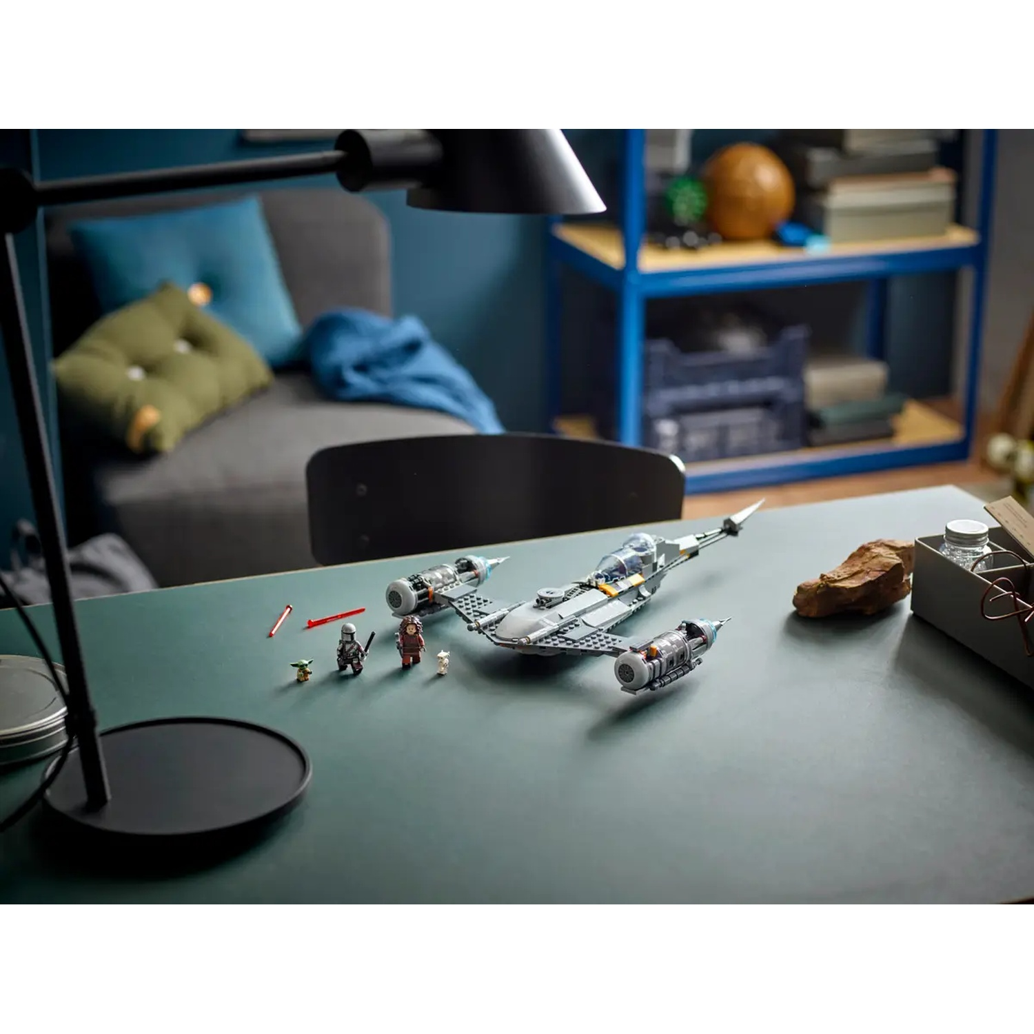 A Lego Star Wars set is on a green desk. It includes a ship and figures. A lamp is in the foreground, and a couch is in the background.